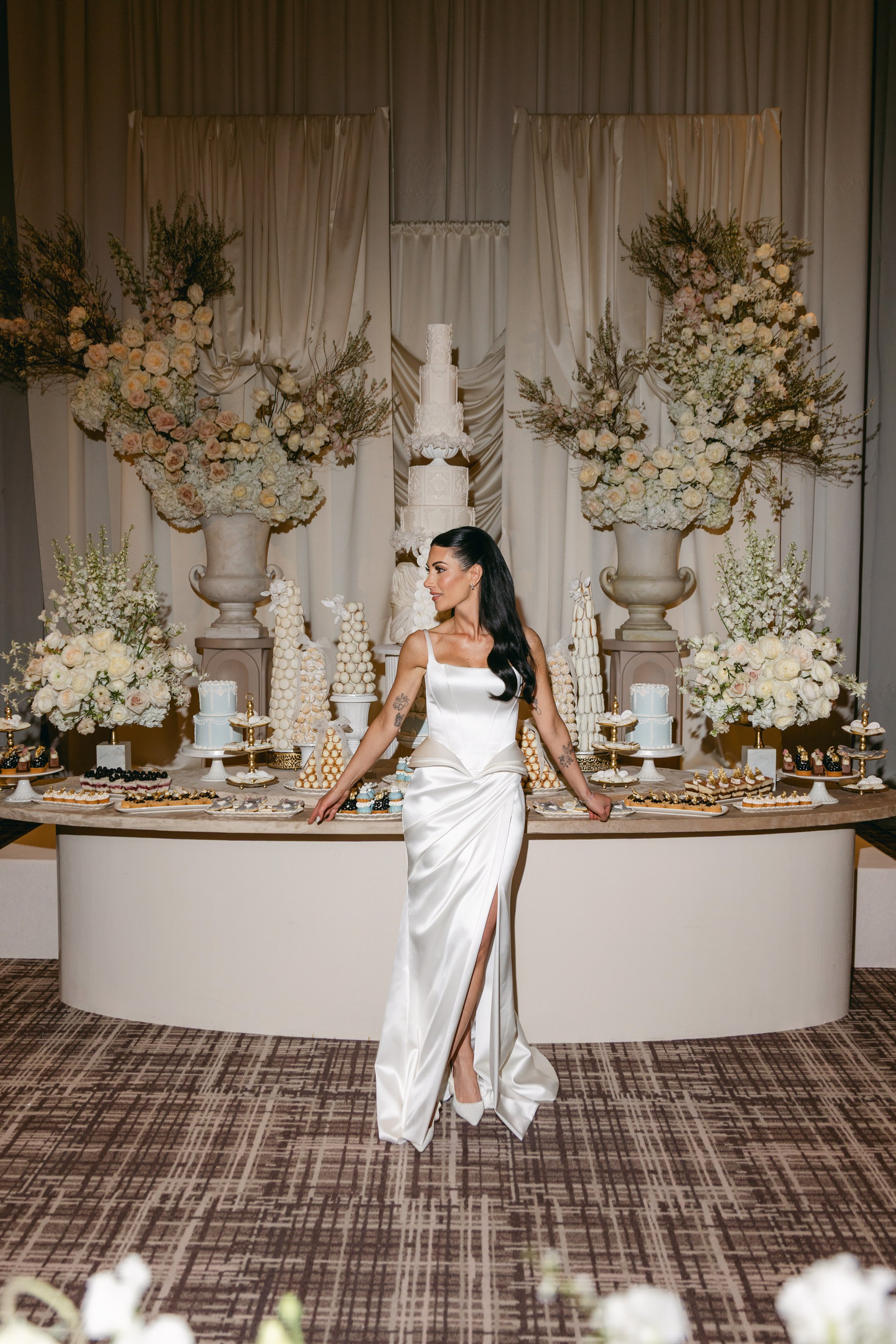 A woman in a white satin dress with a slit standing in front of a dessert table at a wedding or celebration, surrounded by large floral arrangements and a tiered cake.