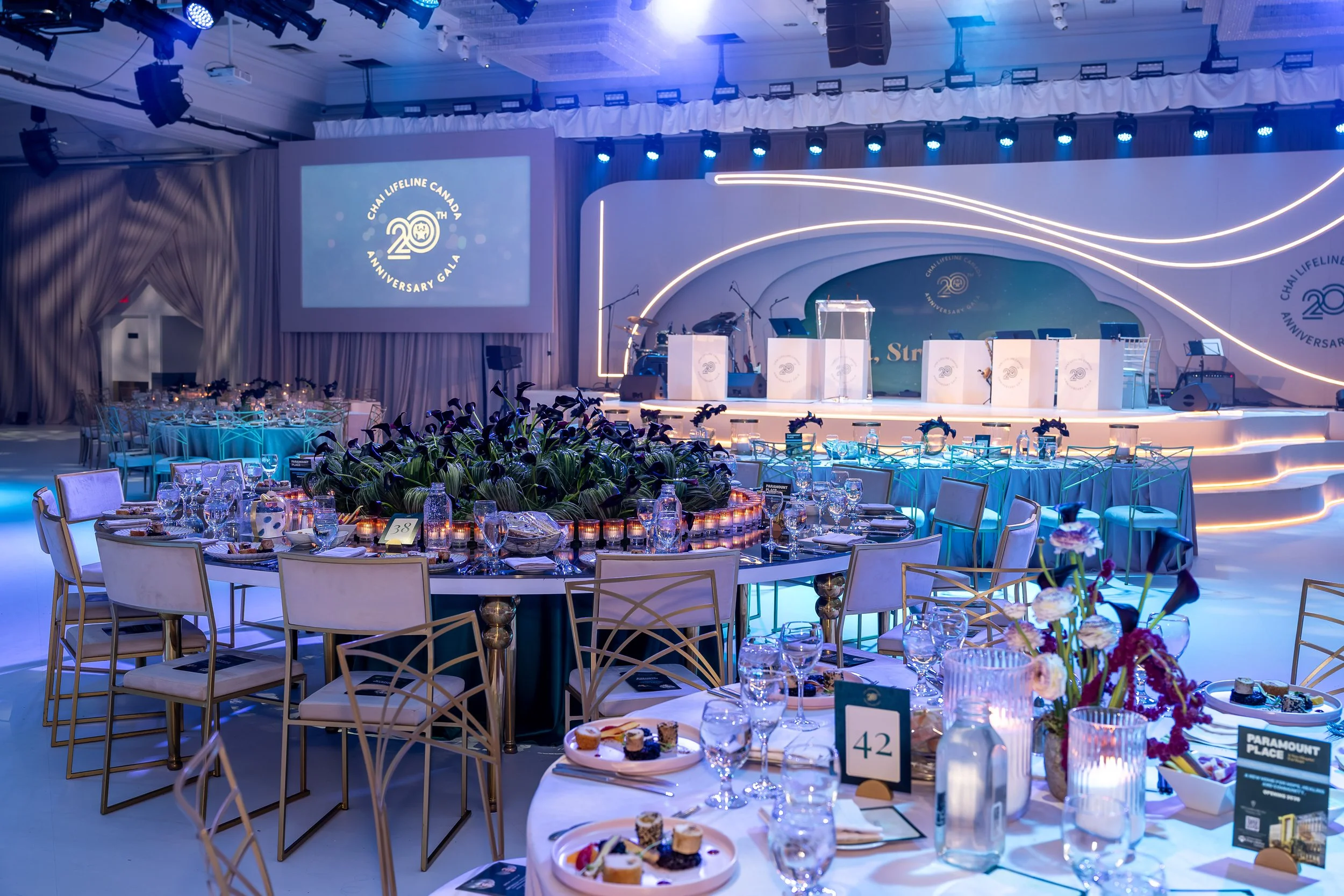 An elegantly decorated banquet hall with round tables set for a celebration, featuring flower arrangements, glassware, and place settings. On the stage, there are musical instruments including drums and microphones, with a large screen displaying the