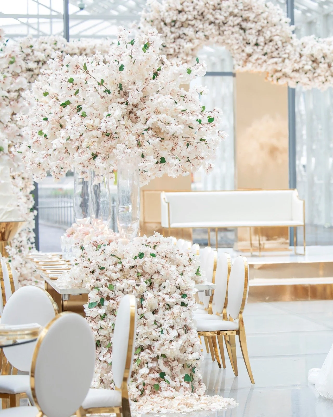 Elegant event setup with large pink floral arrangements, white chairs with gold accents, and a cream-colored bench with a gold base, in a bright, airy space with glass walls.