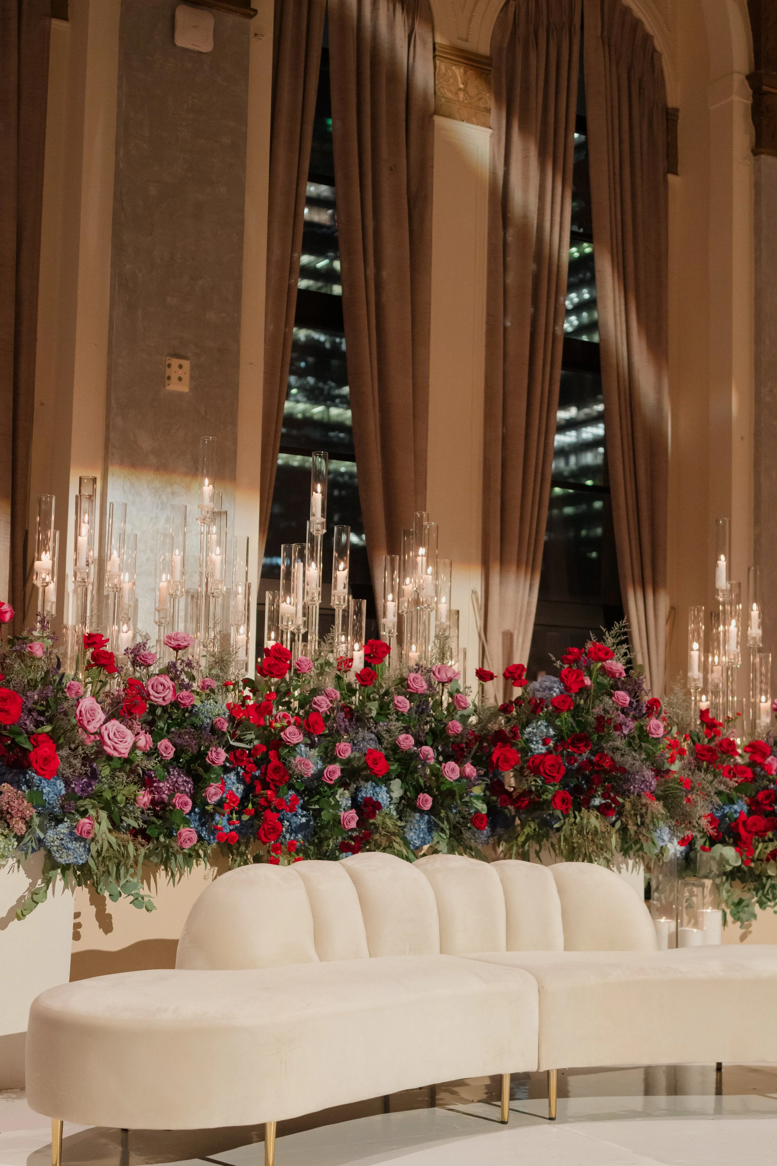 Elegant indoor setting with a cream velvet sofa, a large lush floral arrangement with pink, red, and purple roses, and tall glass candle holders with lit candles, near tall windows with beige curtains.