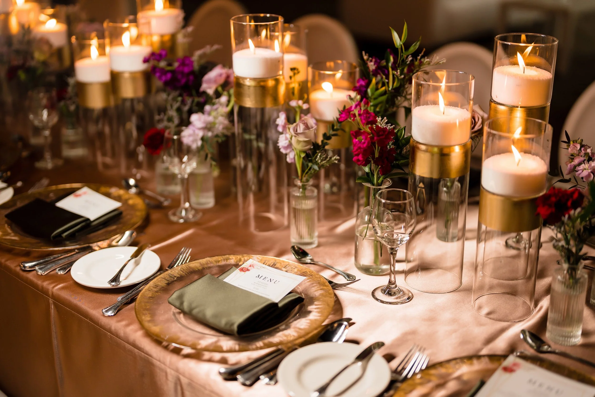 Elegant banquet table decorated with pink satin tablecloth, floral centerpieces with purple, pink, and red flowers, lit candles in glass holders, and place settings with plates, silverware, and menus.