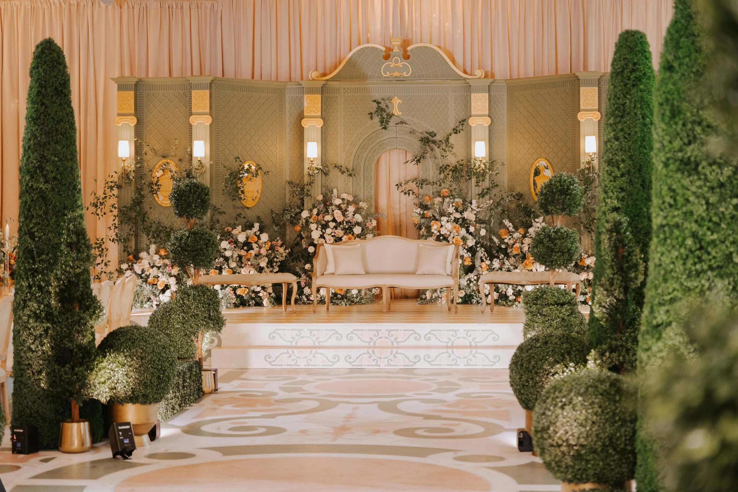 Elegant event stage with cream-colored furniture, surrounded by lush green plants and floral arrangements, with gold accents and soft lighting.