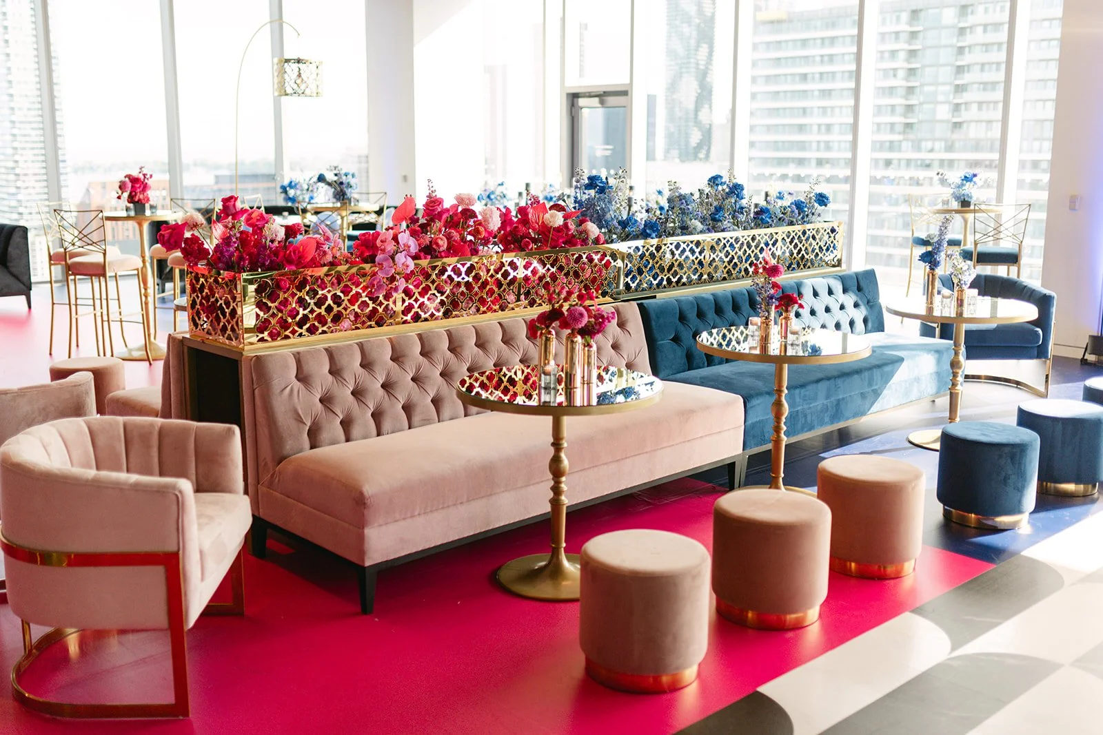 A brightly lit lounge with pink, blue, and beige velvet seating, gold accents, and colorful flower arrangements. Large windows showcase city buildings outside.
