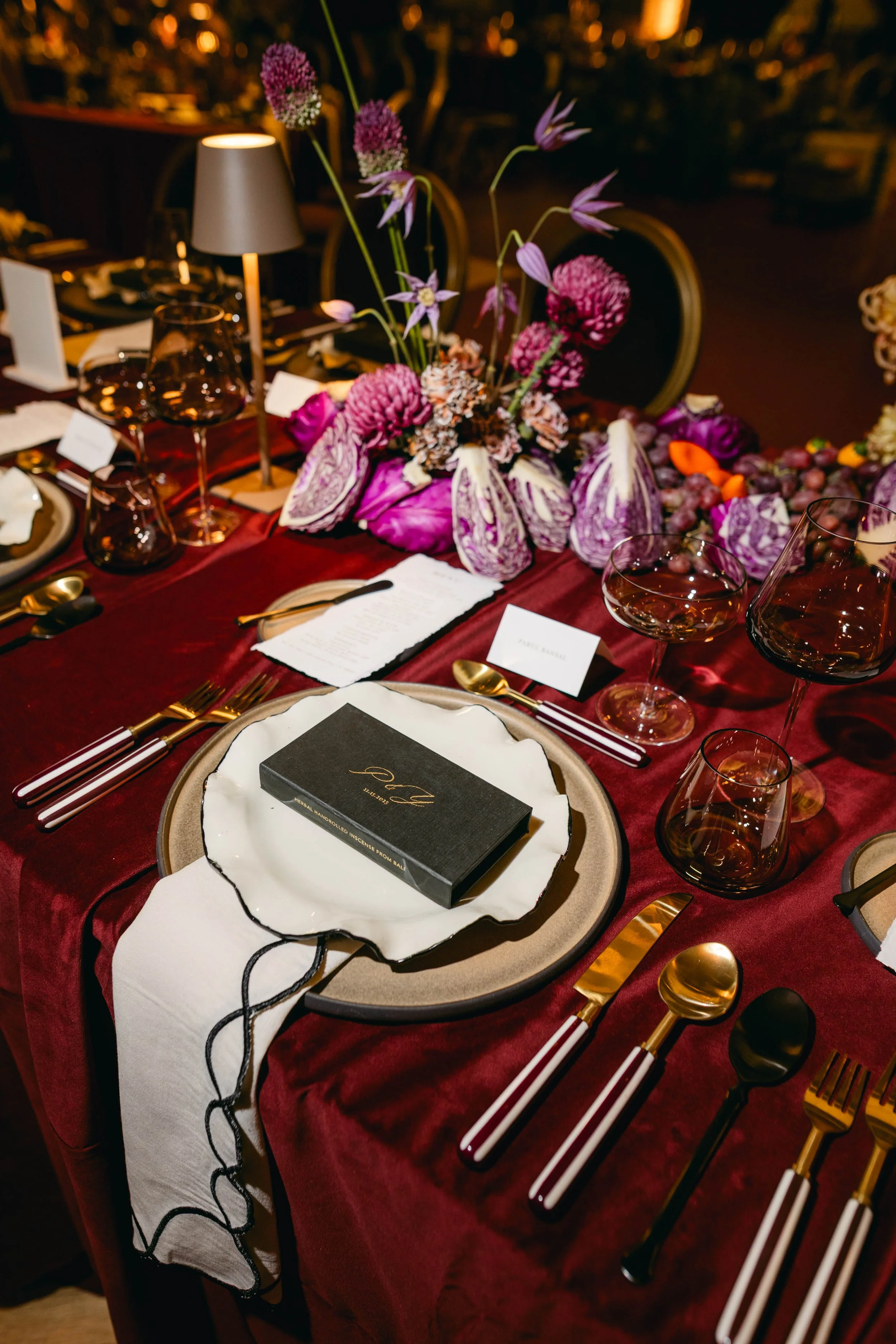 Elegant dining table setting with a large floral centerpiece featuring purple and pink flowers, wine glasses, gold flatware, a menu card, and a black box on a decorative plate, all on a deep red tablecloth and draped white napkin with black embroidery.