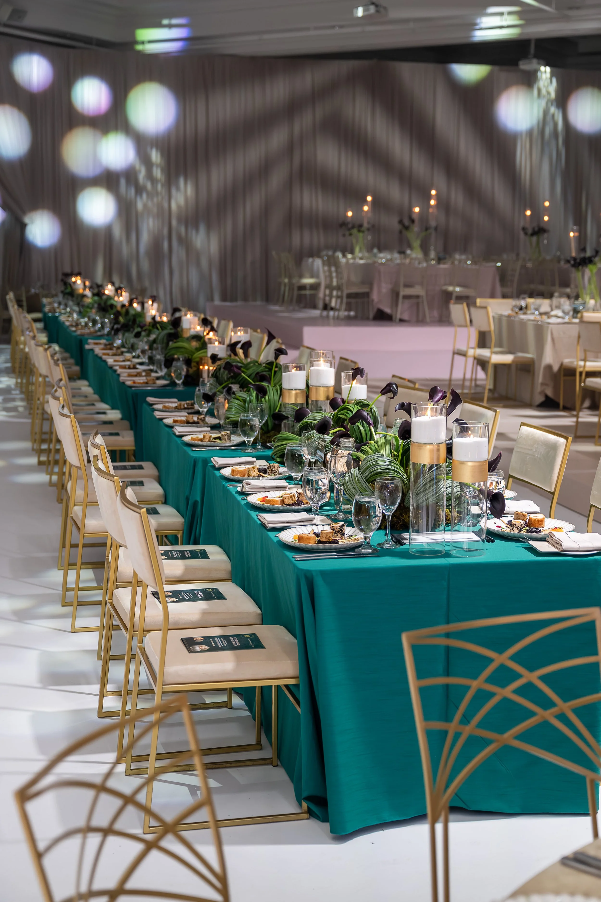 Elegant banquet table set with green tablecloth, floral centerpieces, candles, and place settings for an event inside a decorated room.