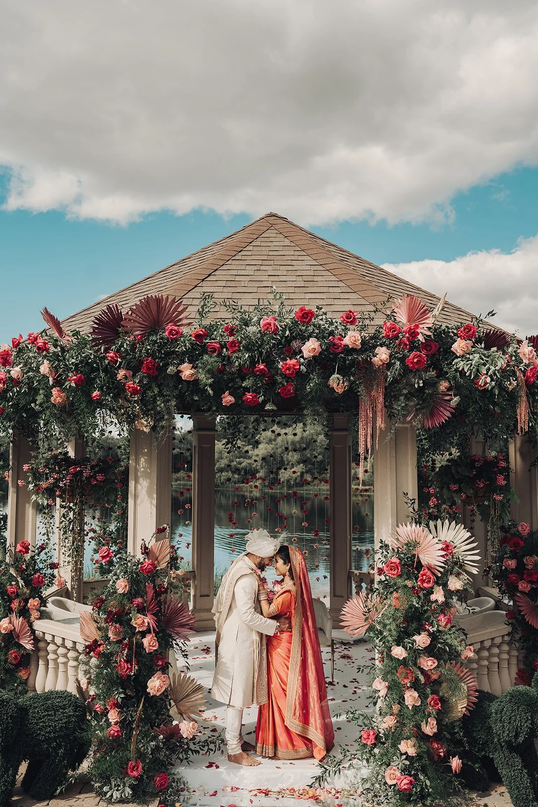 Indian couple on wedding day standing face to face in a decorated outdoor pavilion with flowers, trees, water and sky in background.