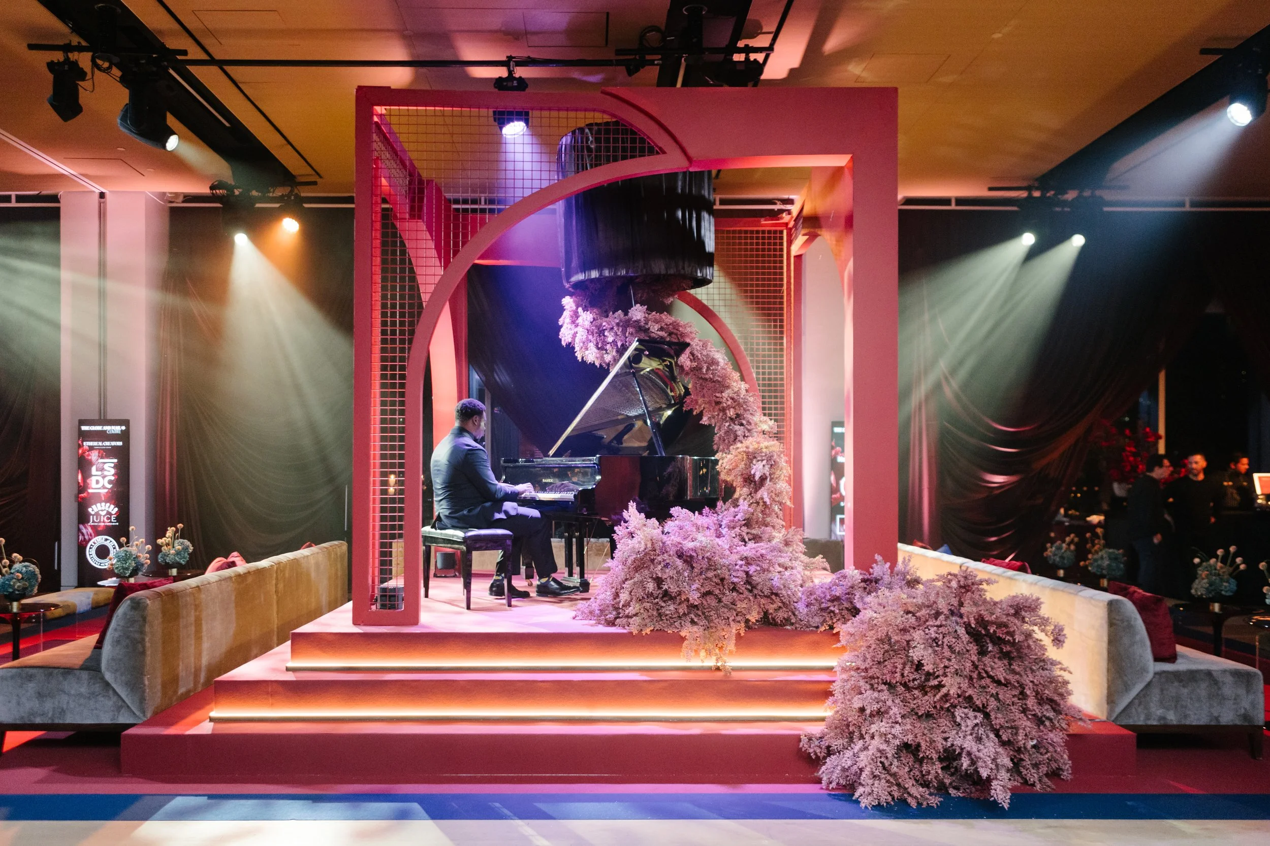 A pianist performing on a decorated stage with pink flowers, illuminated by warm lighting in an indoor event space.