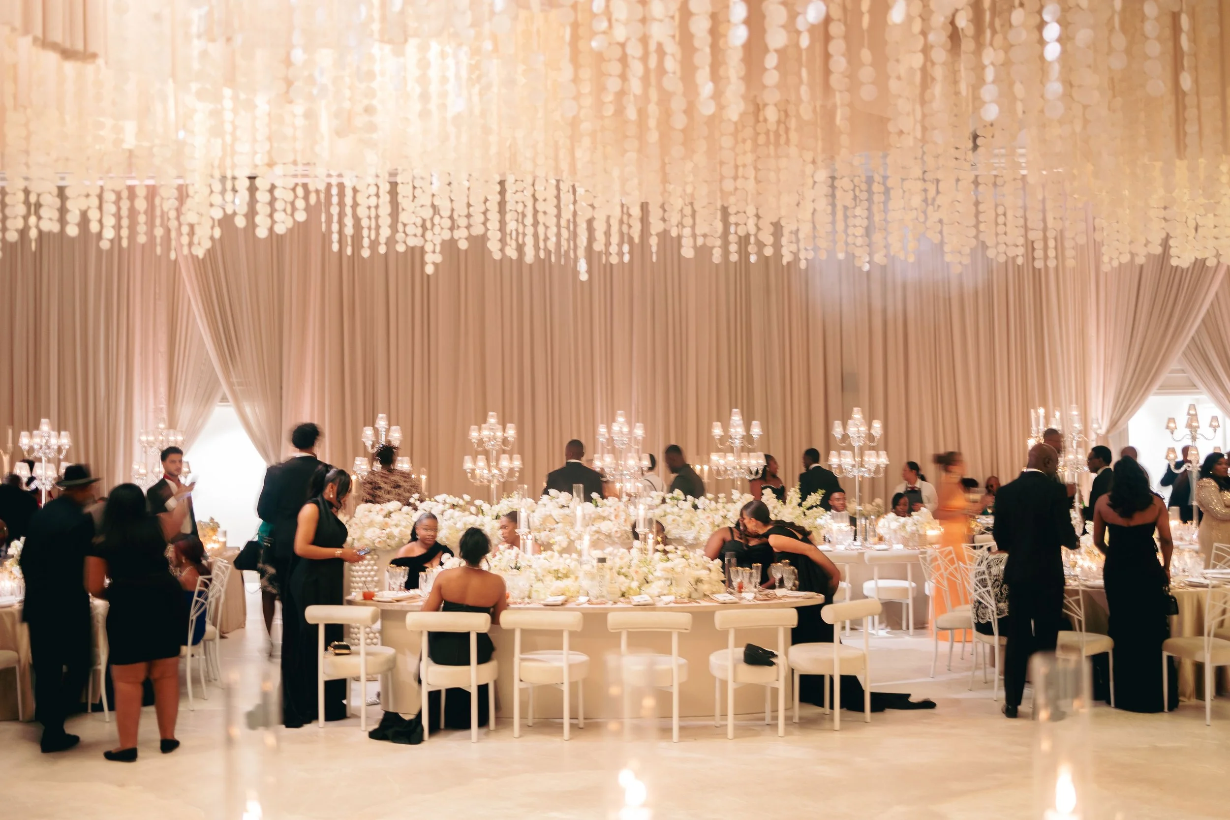 Elegant banquet hall with a high, draped ceiling, filled with people, white tables, and floral centerpieces.