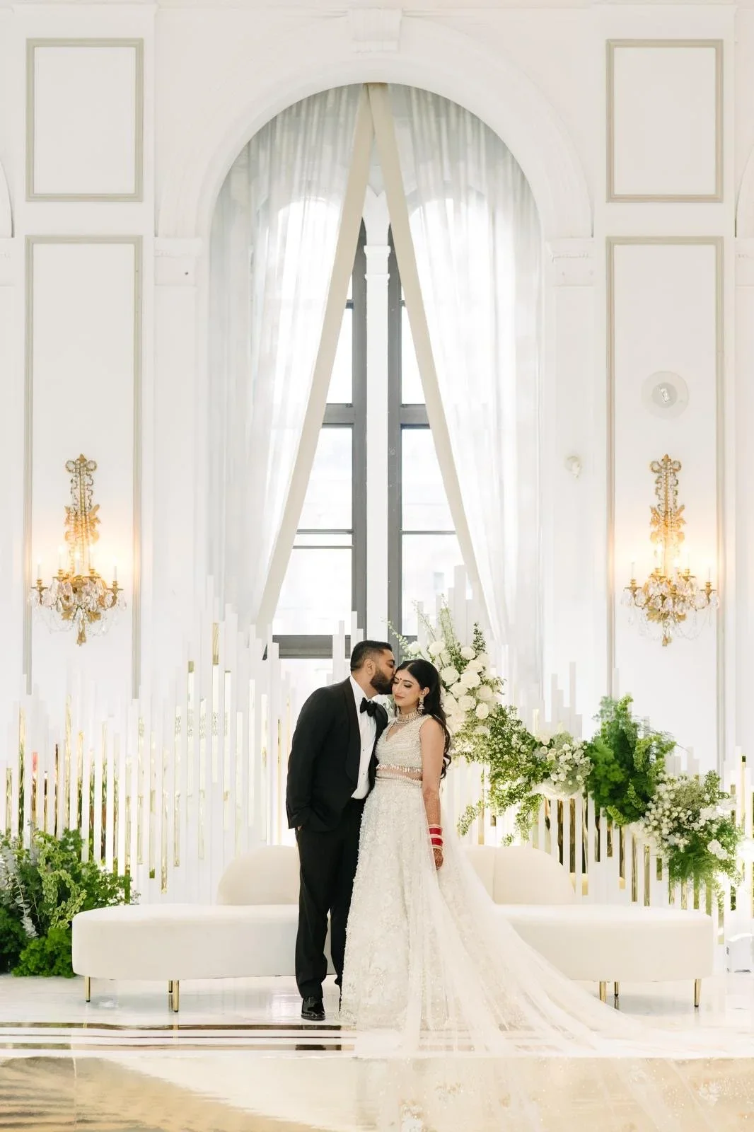 A bride and groom standing close together at their wedding in an elegant, bright white venue with large windows, white curtains, floral arrangements, and ornate wall fixtures.