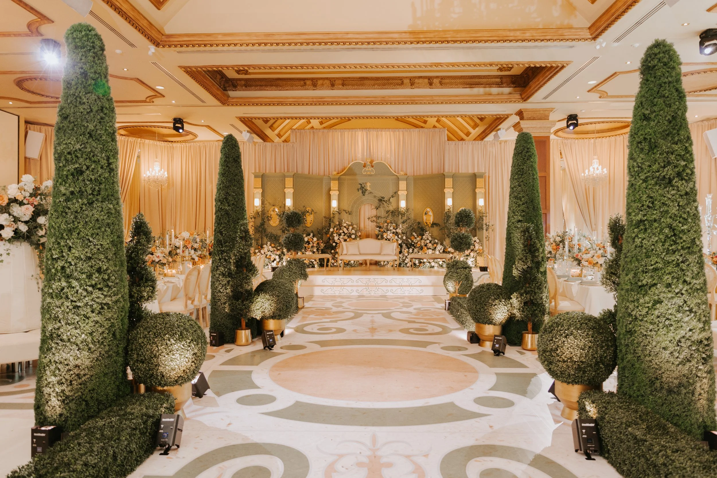 Elegant event space featuring a stage with a white sofa, surrounded by floral arrangements and decorated with greenery, tall cypress trees, and soft lighting, suitable for a wedding or formal celebration.
