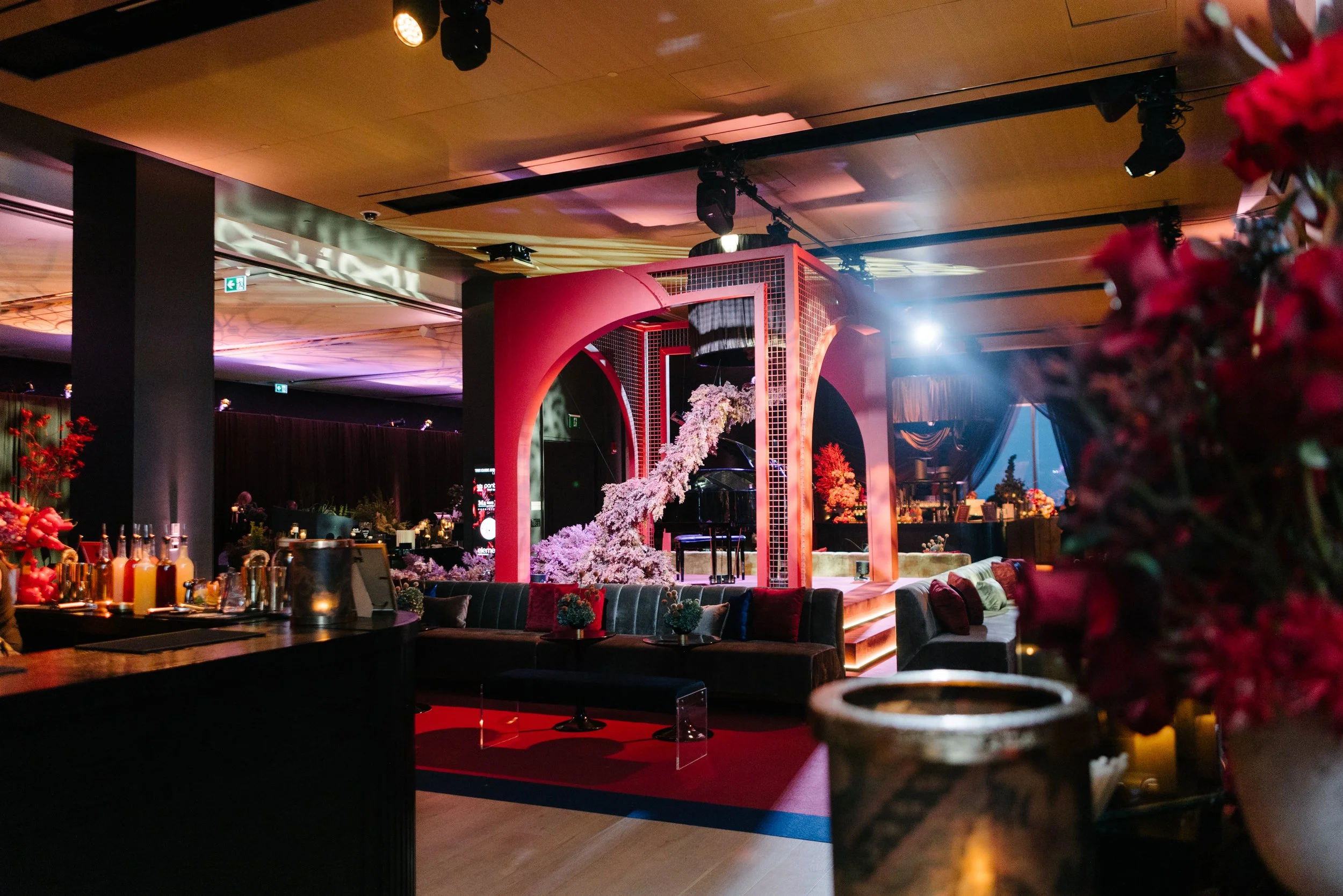 An elegantly decorated indoor event space with a stage featuring pink floral arrangements and a grand piano, surrounded by cozy seating and ambient lighting.