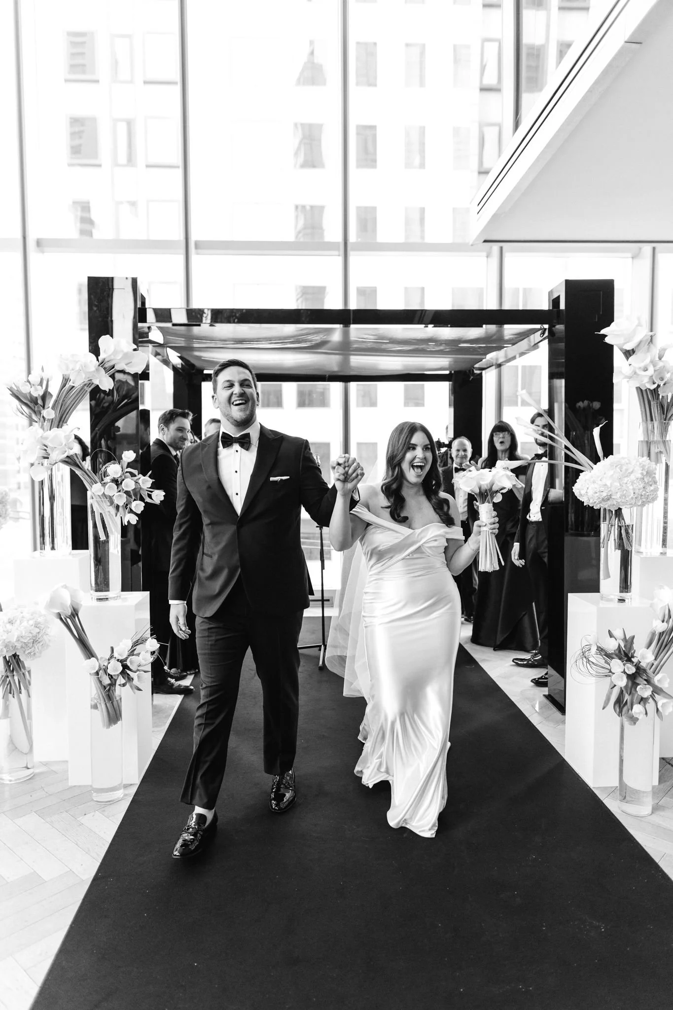 A newlywed couple walking hand in hand down the aisle, smiling. The groom wears a tuxedo and the bride wears a satin wedding dress, holding a bouquet. They are in a modern, glass-walled interior with floral decorations.