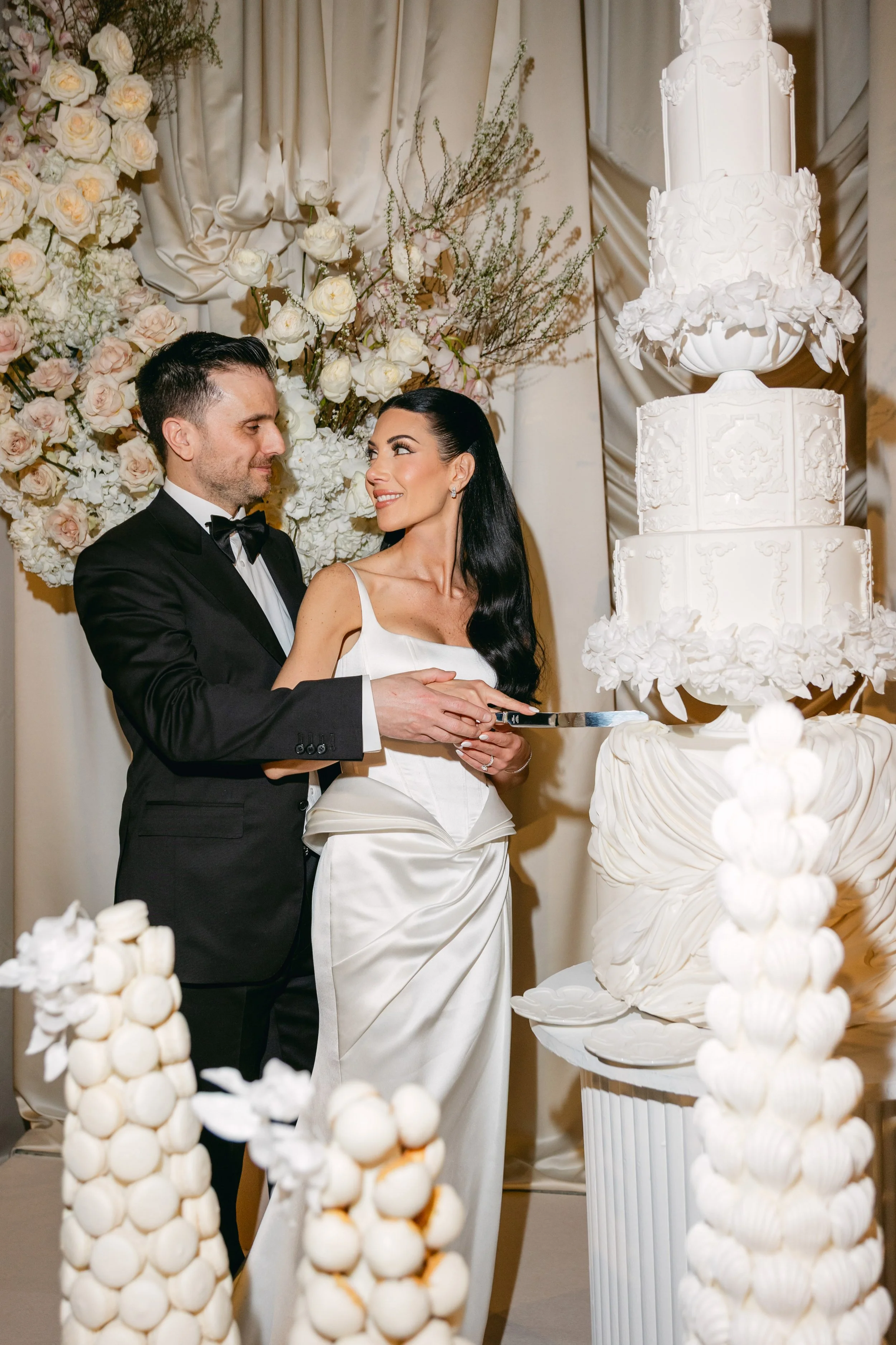 A bride and groom cutting a white wedding cake with multiple tiers, surrounded by white floral decorations and cream-colored drapes.