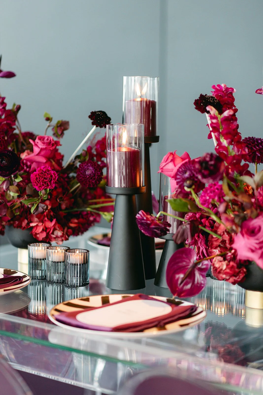 Table decorated with pink and purple flowers, tall red candles in glass holders, and dark-colored plates with napkins.