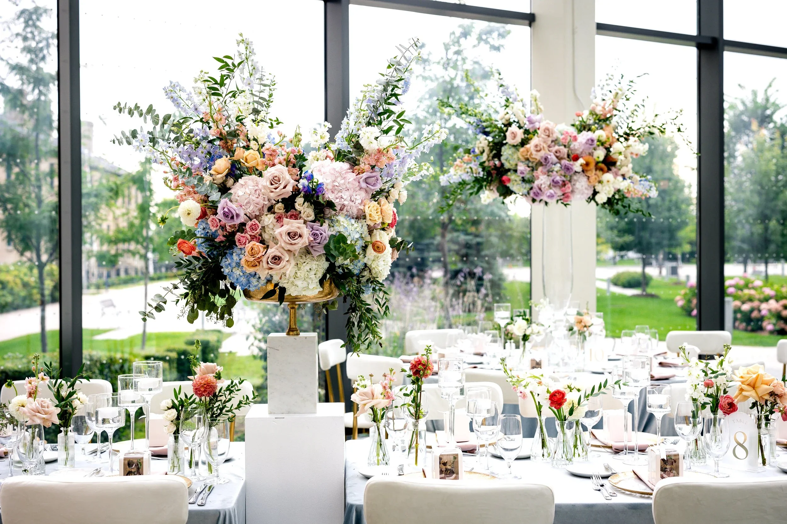 Elegant banquet table setup in a bright, modern room with large windows overlooking a green outdoor landscape, featuring tall floral centerpieces with pastel roses, hydrangeas, and greenery, and neatly arranged glassware, plates, and utensils.