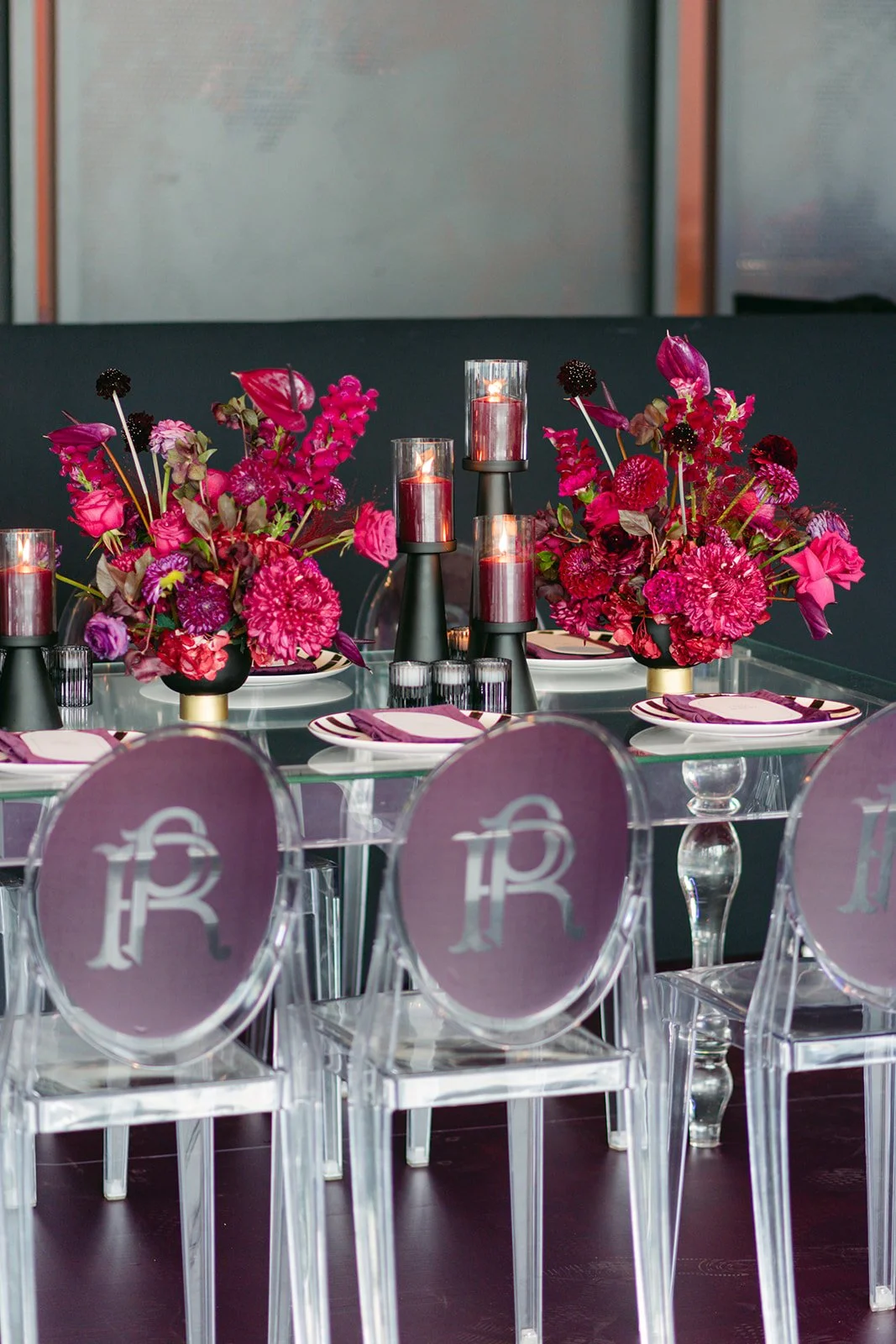 Elegant table setting with vibrant pink floral centerpieces, lit pink candles in black holders, and clear acrylic chairs with monogrammed backs.