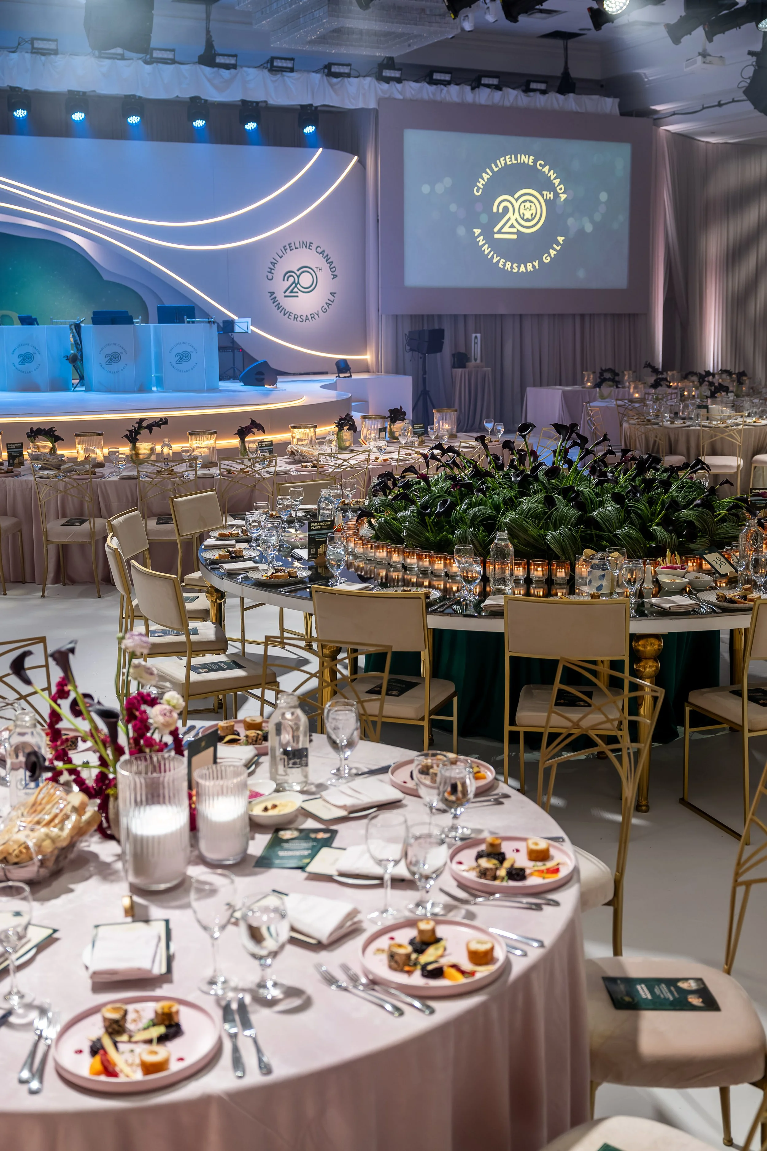  banquet hall decorated for a formal celebration, with round tables set with plates, glassware, and floral centerpieces, and a stage with a large screen displaying the 20th anniversary of Chai Lifeline Canada.
