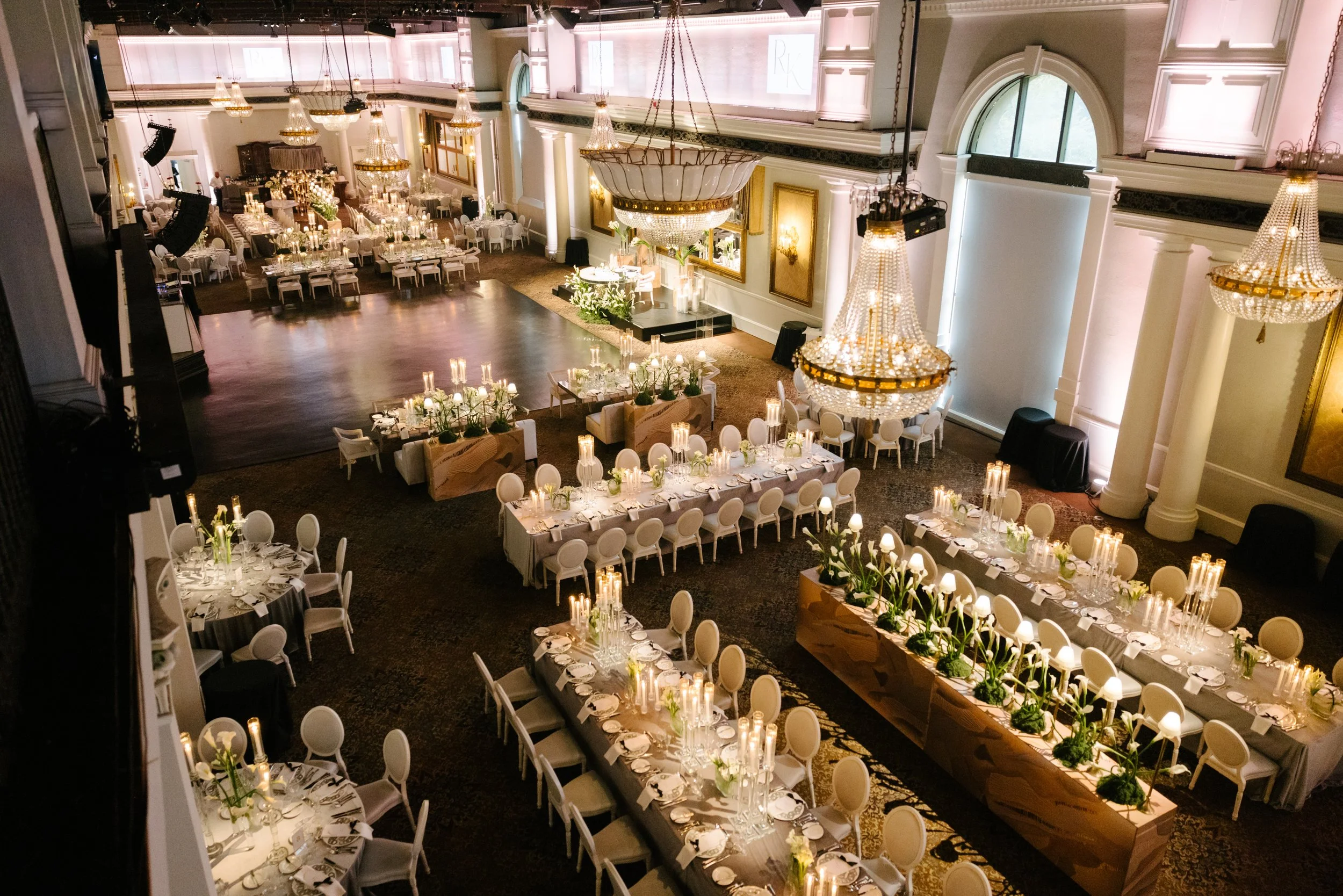 Elegant banquet hall decorated with large chandeliers, long dining tables with white chairs, floral centerpieces, and candles, set for a formal event or celebration.