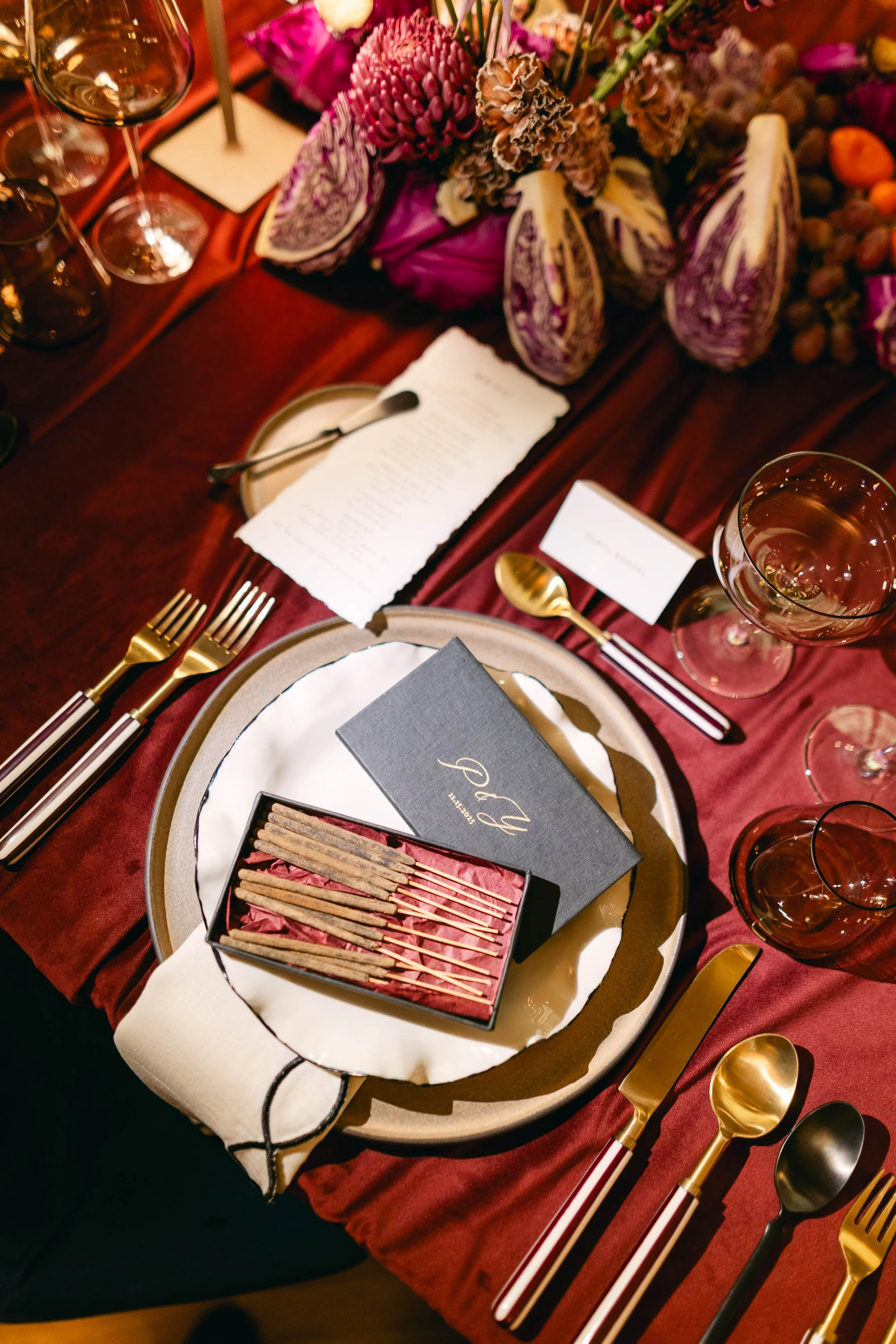 A formal dining table setting with wine glasses, gold and black cutlery, a menu, a gift box with matchsticks, a notebook, and a floral centerpiece.