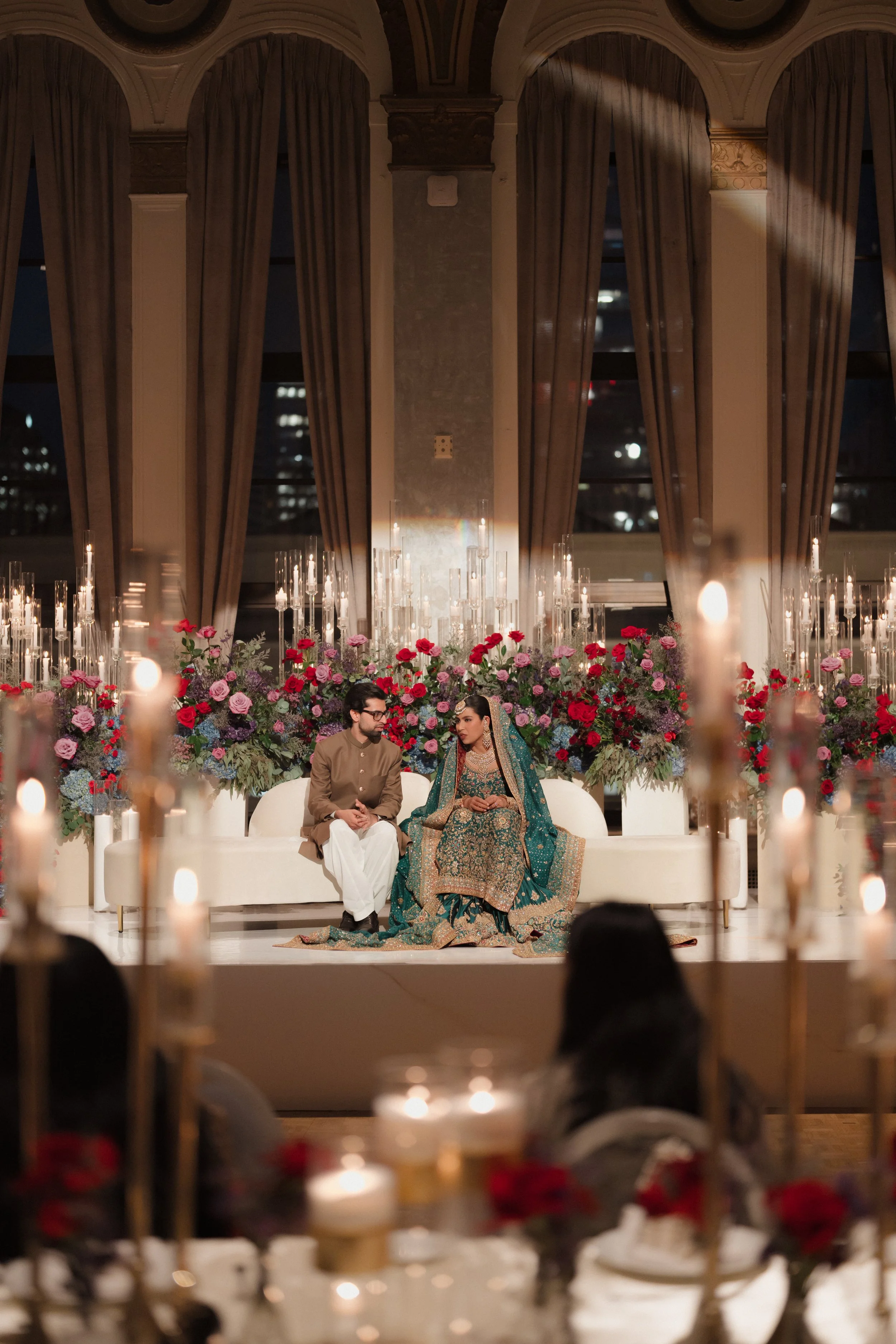 A bride and groom seated on a white sofa during a wedding ceremony, surrounded by a floral arrangement and candles, inside a decorated hall with tall curtains and large windows at night.
