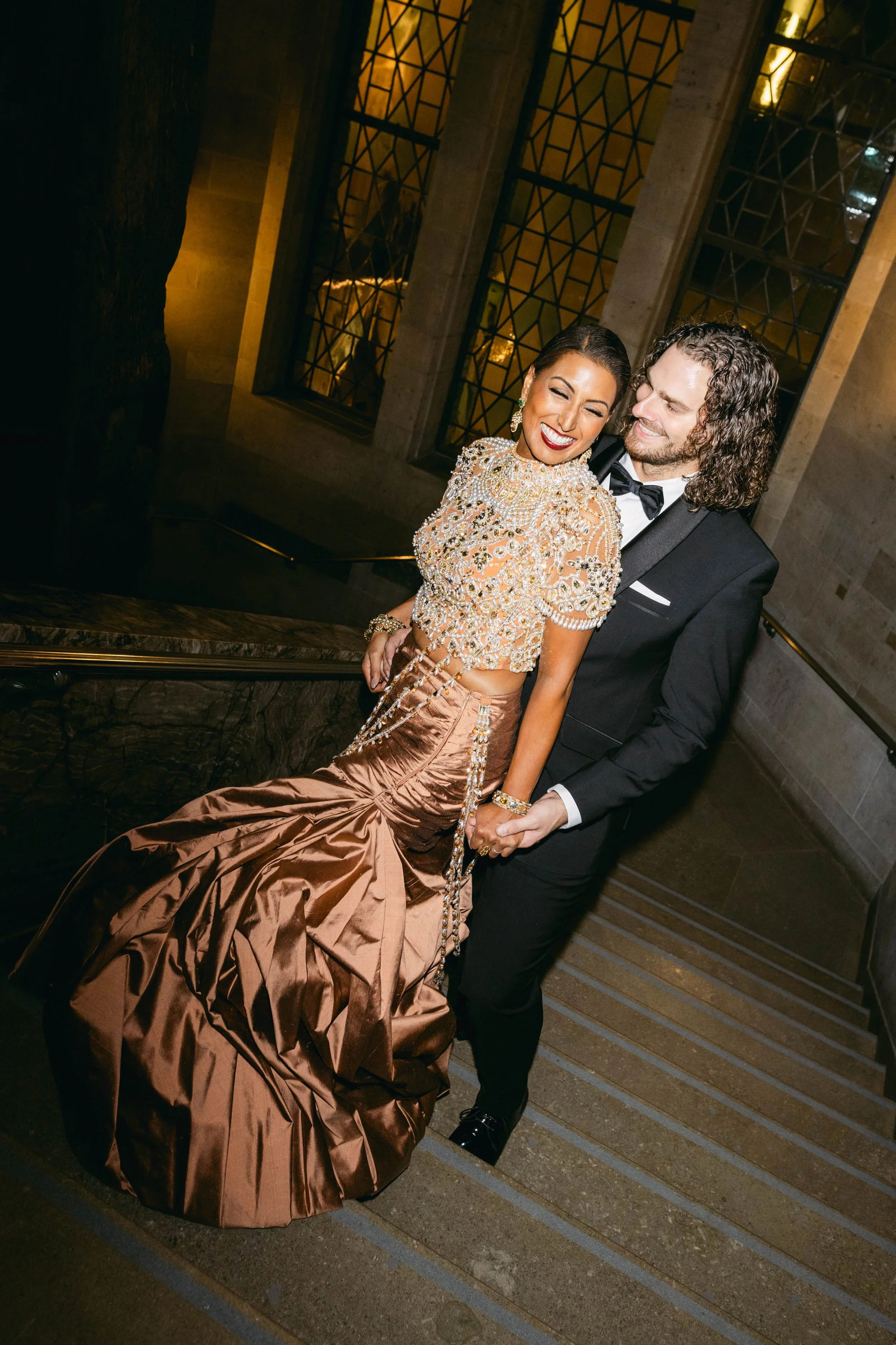 A smiling woman in an ornate, beaded top and bronze satin skirt, with a man in a tuxedo, holding her hand as they descend a staircase at an elegant event.
