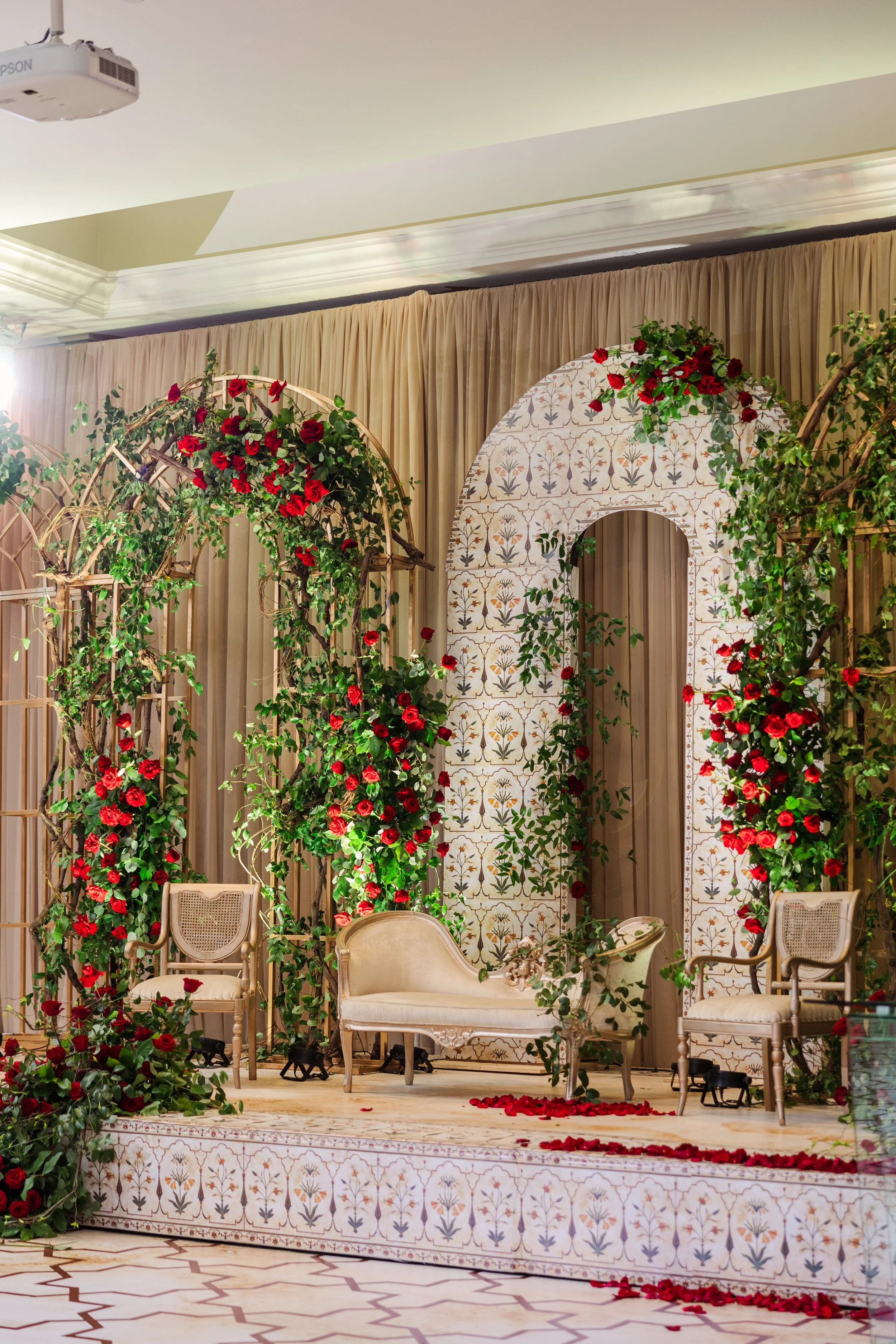 Decorated wedding or event stage with beige curtains, floral arch with red roses and green foliage, vintage chairs, and a white ornate bench, set on a decorated platform with scattered rose petals.