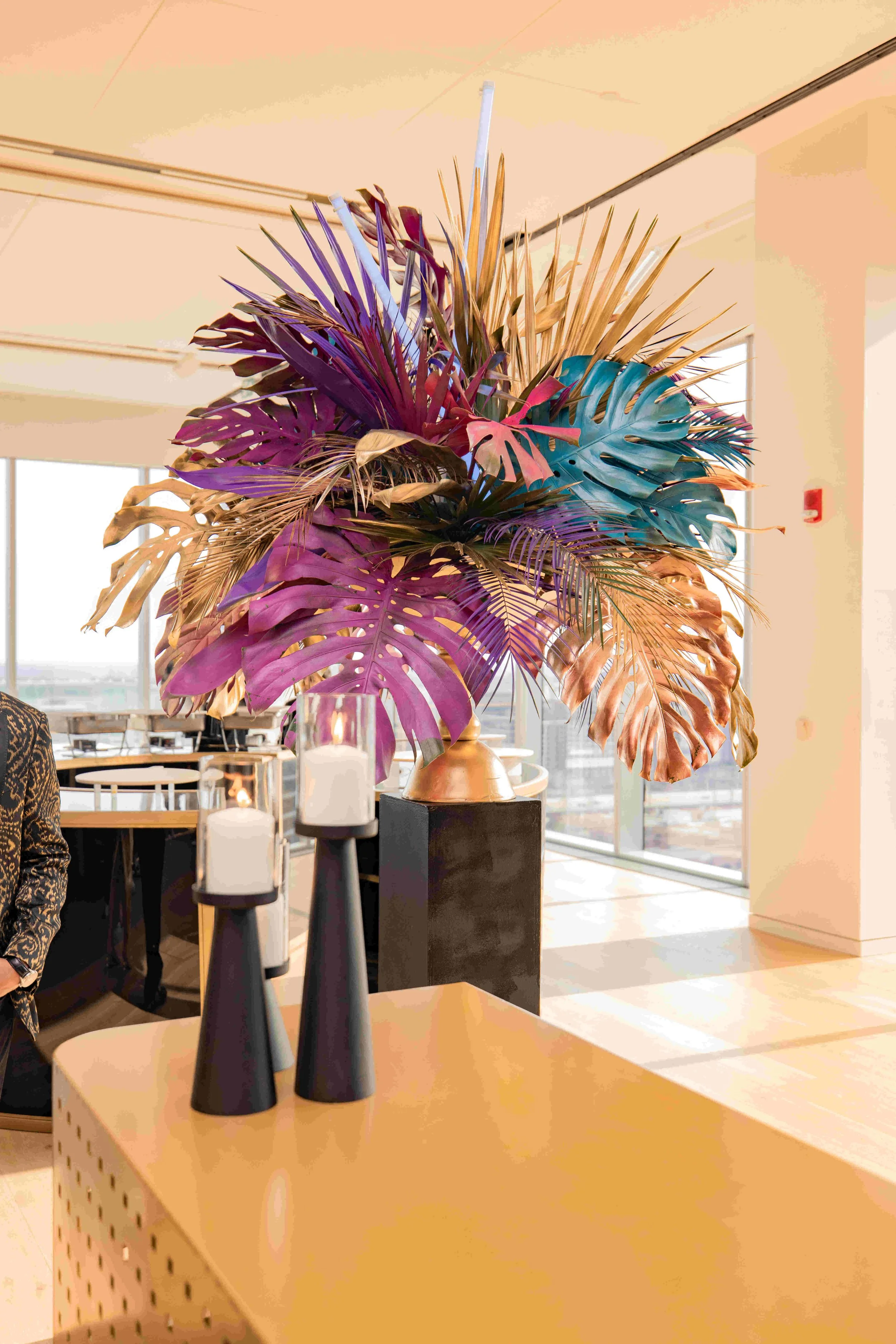 A large, colorful floral arrangement with various leaves in purple, pink, teal, gold, and tan tones, placed in a gold vase on a black stand in a modern indoor space with natural light.