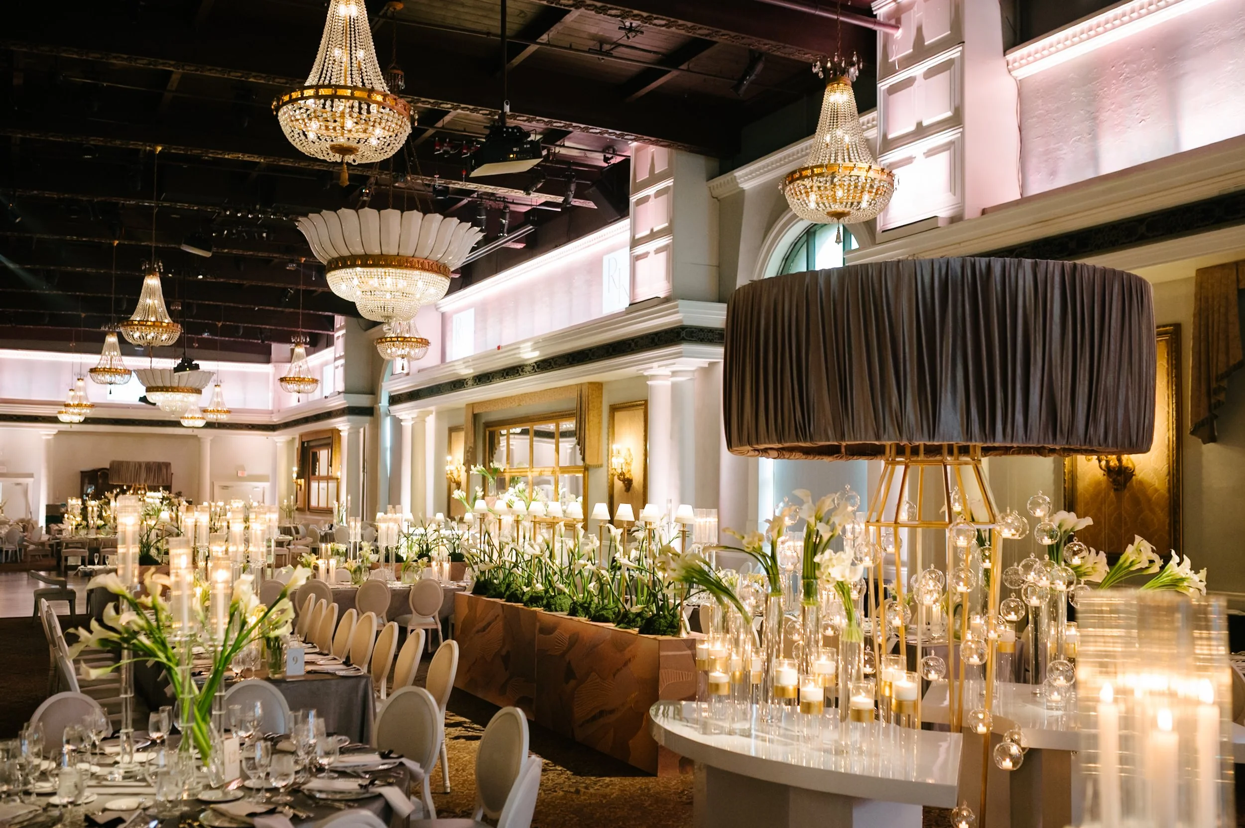 Elegant banquet hall decorated with chandeliers, flowers, candles, and neatly arranged tables for a formal event.