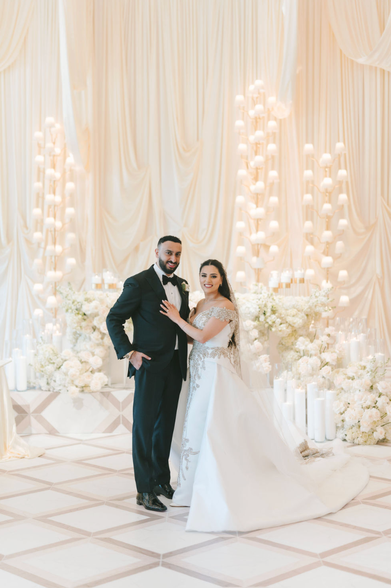 Bride and groom in wedding attire standing in front of a decorated backdrop with white flowers and candles.