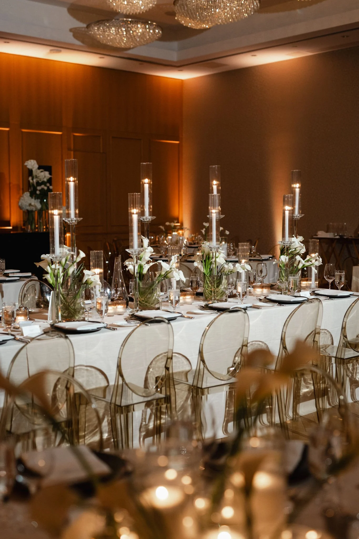Elegant banquet room with decorated tables, tall candles in glass holders, and floral centerpieces, illuminated by warm lighting and chandeliers.