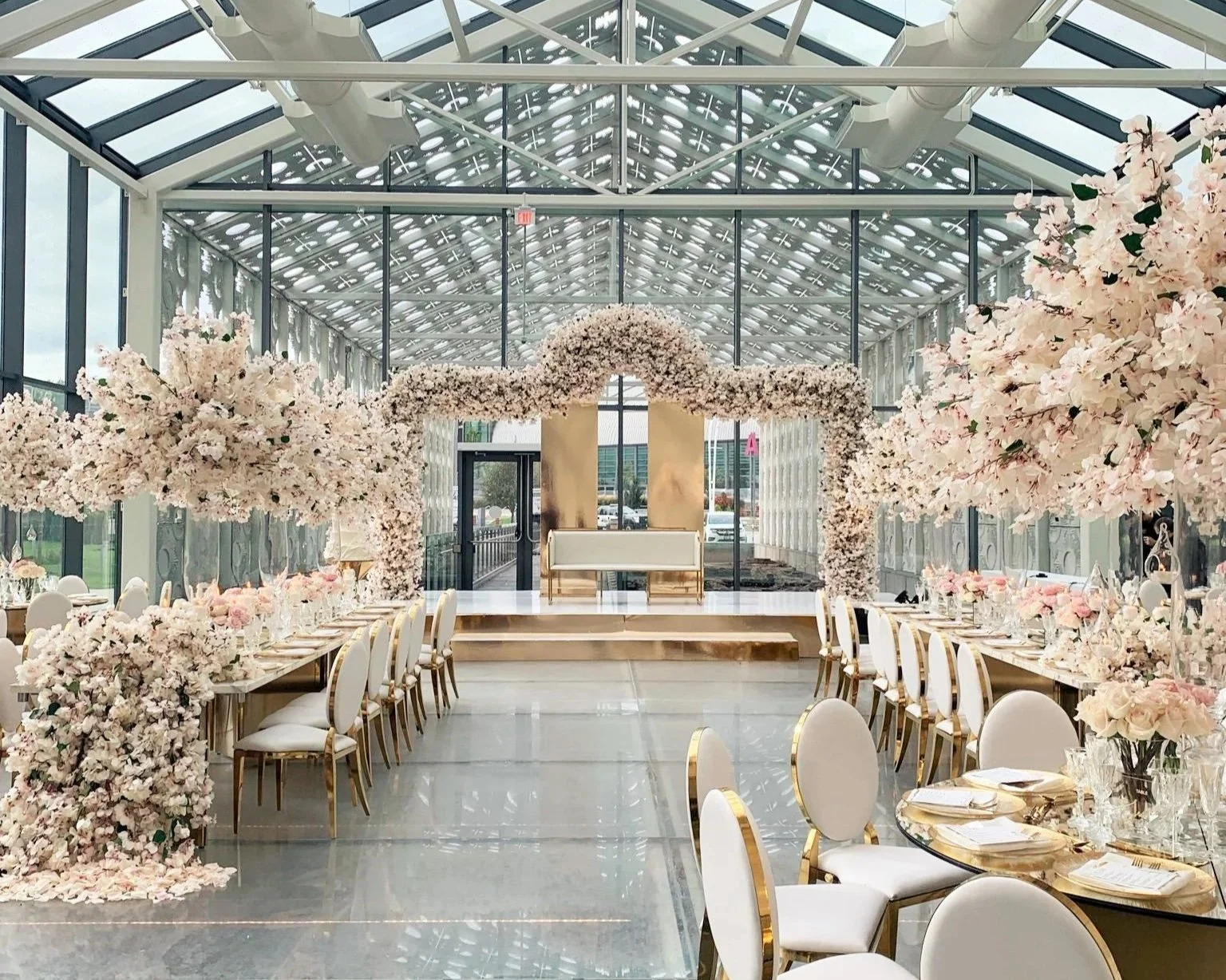 A large, elegant indoor wedding reception venue with glass ceiling and walls, decorated with abundant pink and white floral arrangements, a gold and white stage with a bench, and round tables set with fine china and floral centerpieces.