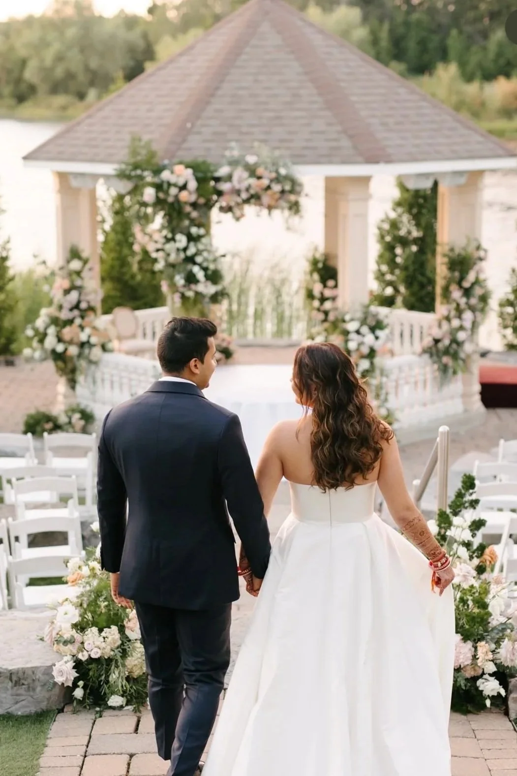Couple walking hand in hand to a wedding altar outdoors, decorated with flowers and greenery, with a gazebo in the background.