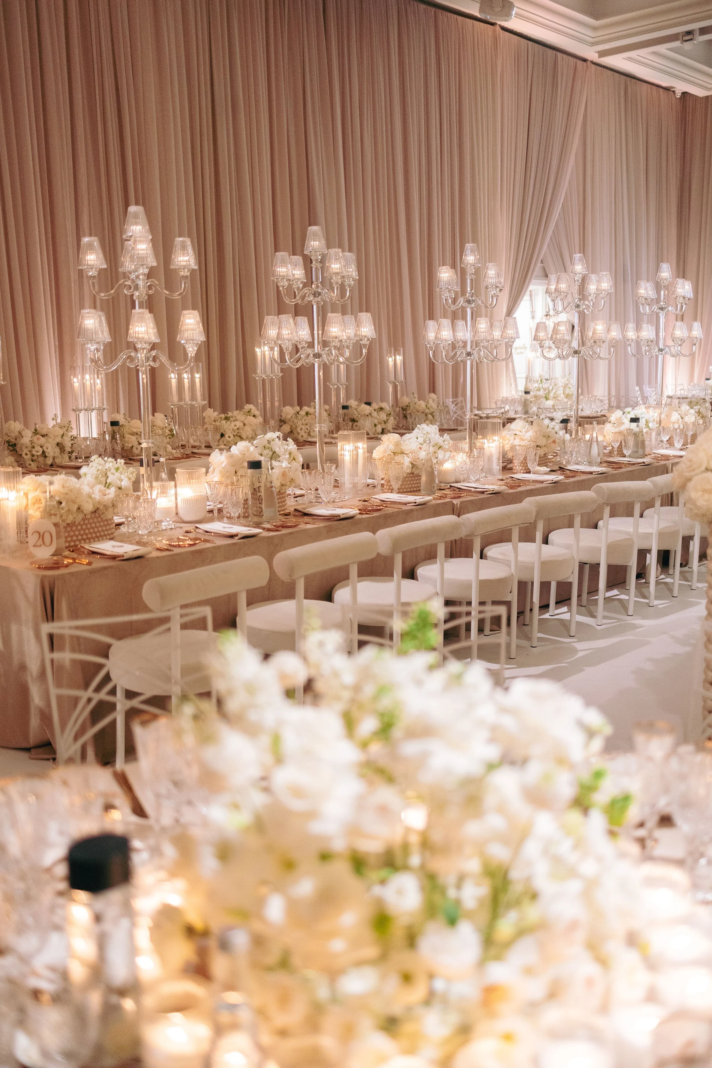 Elegant banquet table decorated with white flowers, candles, and chandeliers in a softly lit room with beige curtains.