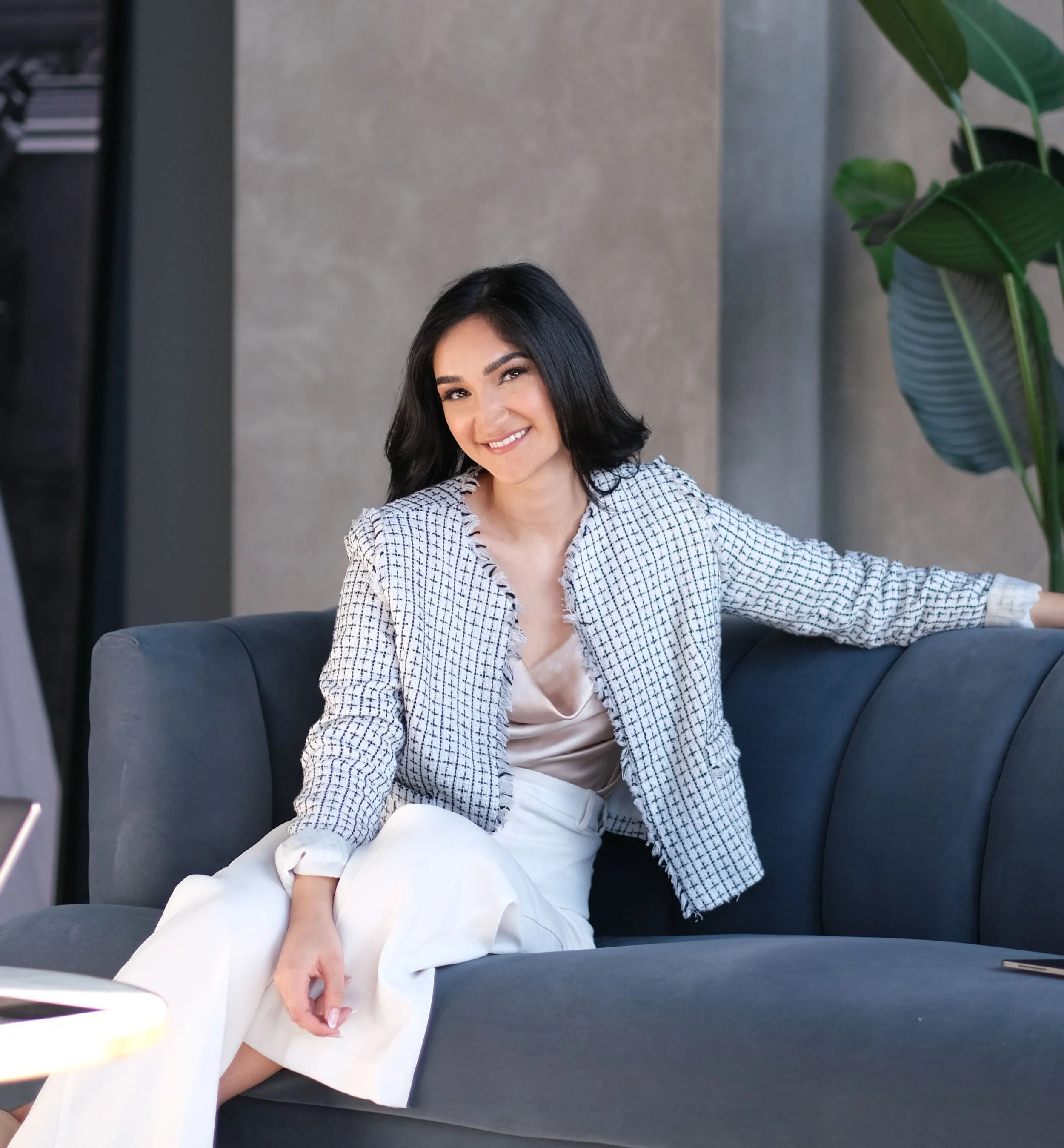 A woman with black hair and a smile, sitting on a gray sofa in a modern, minimalistic room with a concrete wall and a large green plant beside her.
