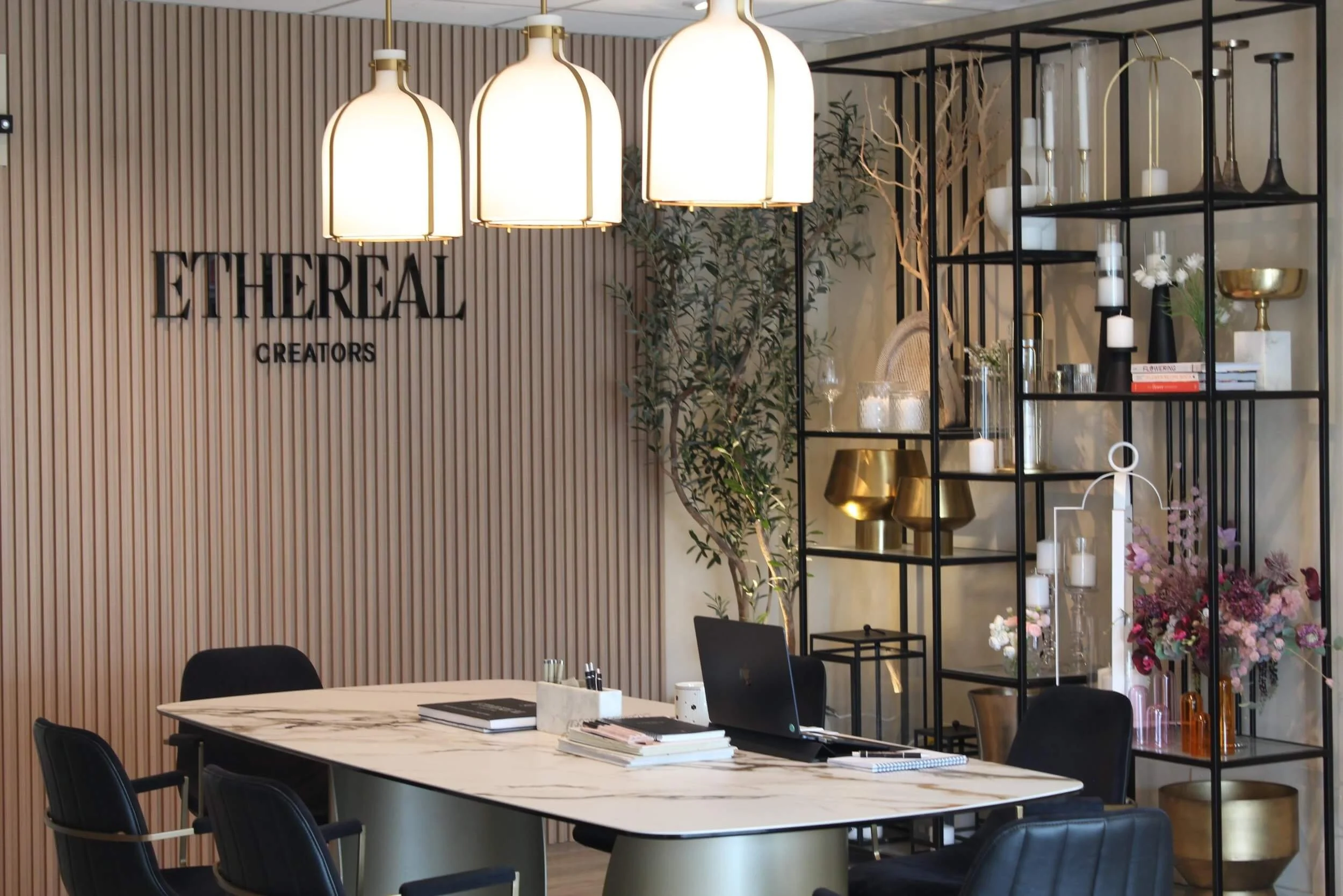 Modern office space with a marble table, black chairs, decorative shelving with gold and white accessories, and a wall with the text 'ETHEREAL CREATORS'