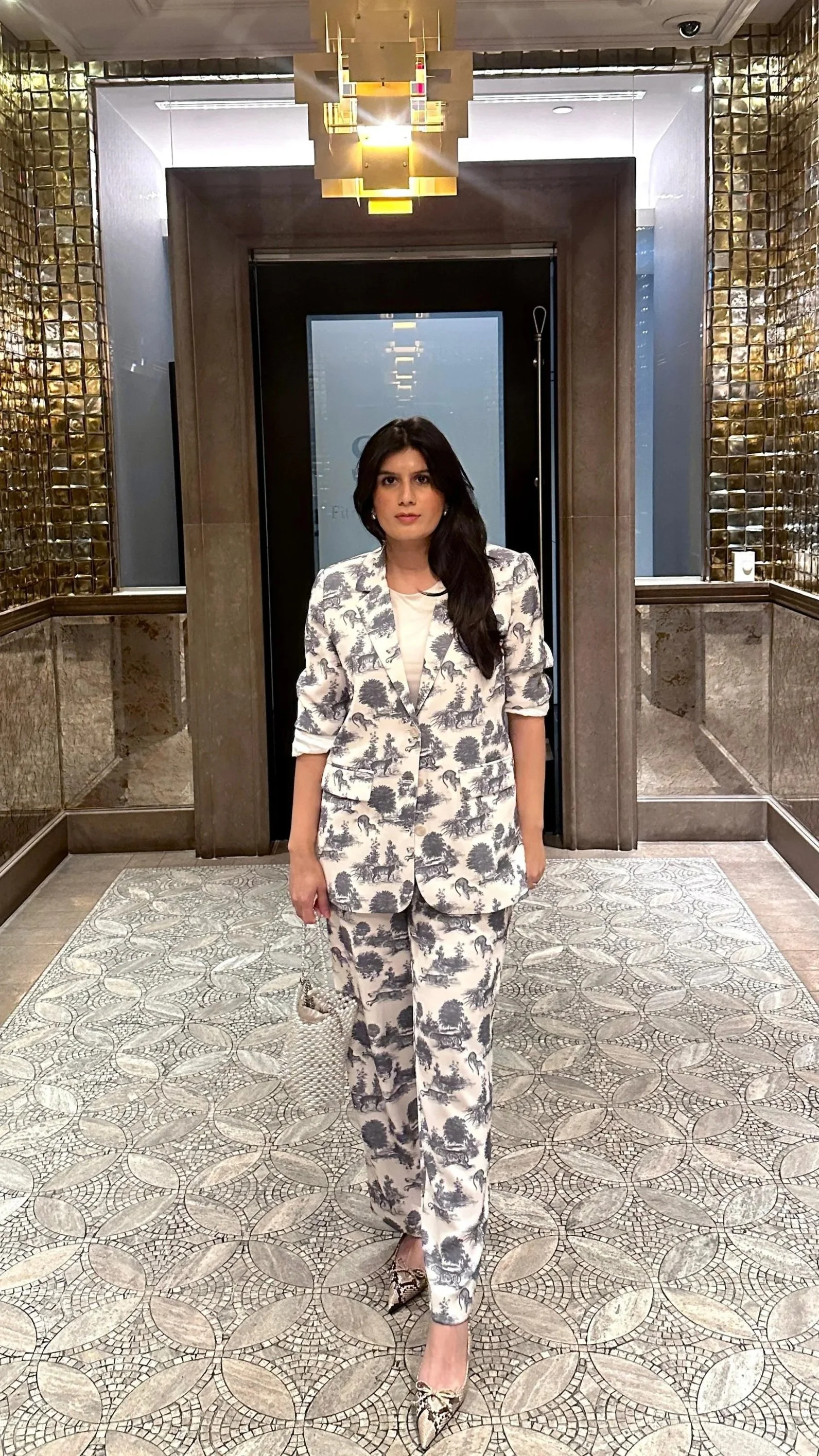 A woman in a matching black and white toile print suit standing in an ornate hotel lobby with gold accents and patterned tile floor.