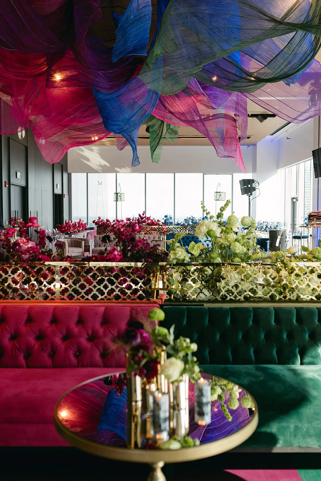 Colorful interior of a decorated event space with hanging fabric art, floral arrangements, velvet seating, and large windows.