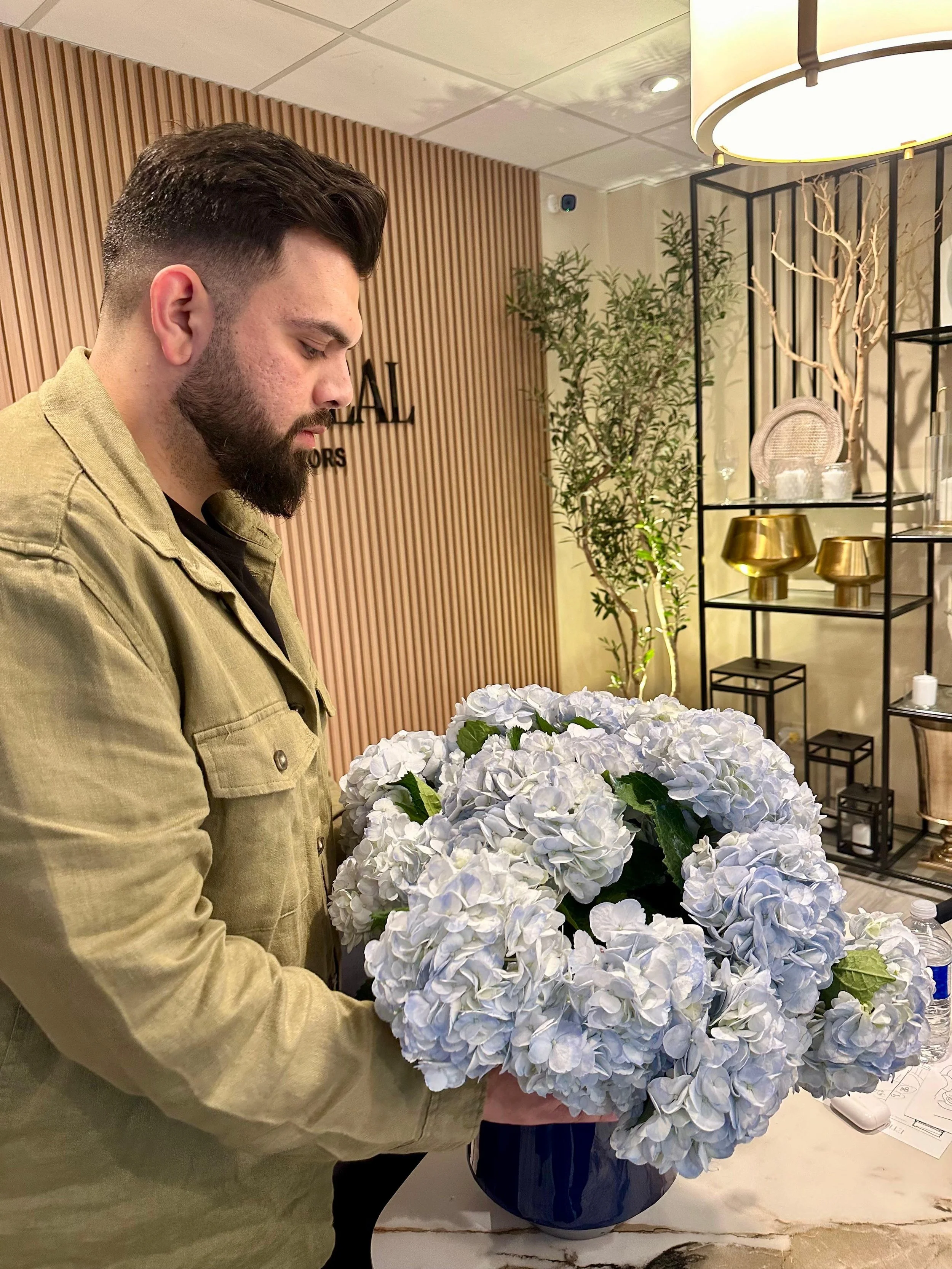 A man with a beard and dark hair is holding a large bouquet of white hydrangeas in a dark blue vase. The background features a wooden slat accent wall with decorative items and a plant.