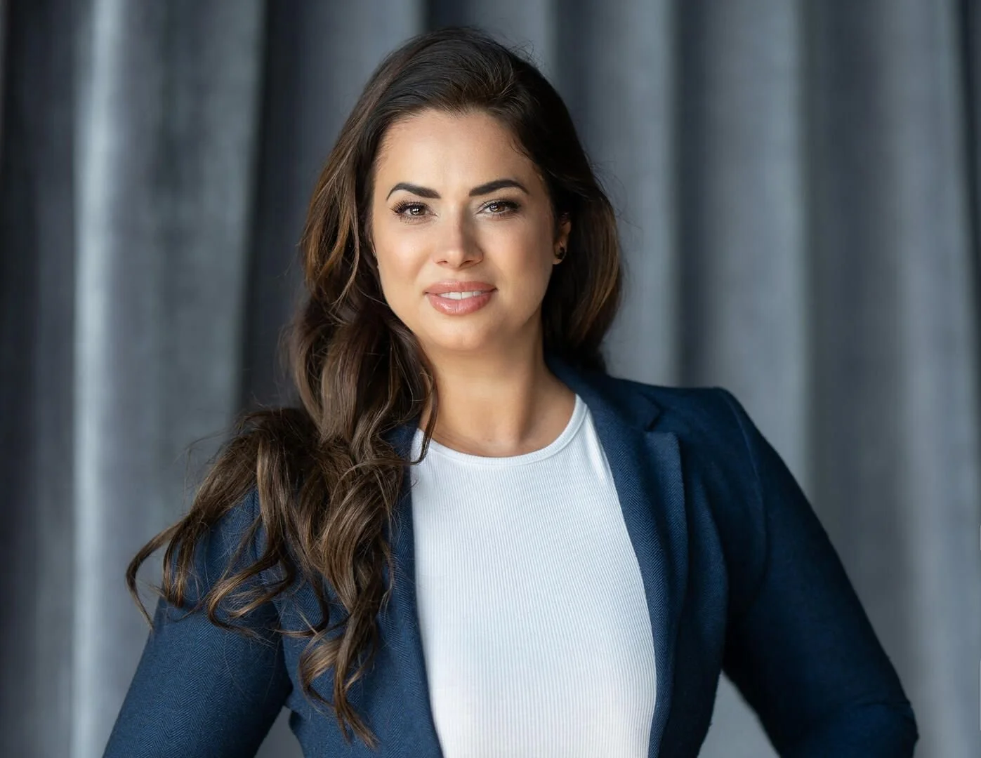 A woman with long, wavy brown hair and a confident expression wearing a white top and a dark blazer, standing in front of gray curtains.