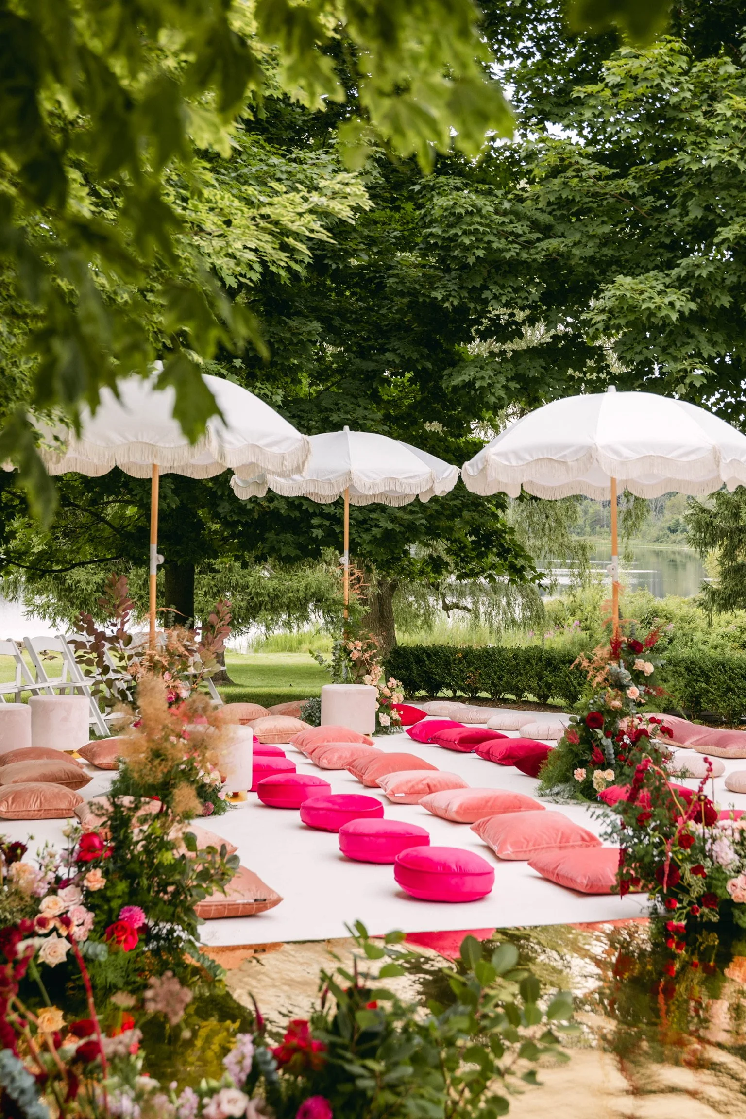 Outdoor setting with large white umbrellas, pink and peach cushions arranged on a white mat under the trees, floral arrangements surrounding the area, near a body of water in the background.