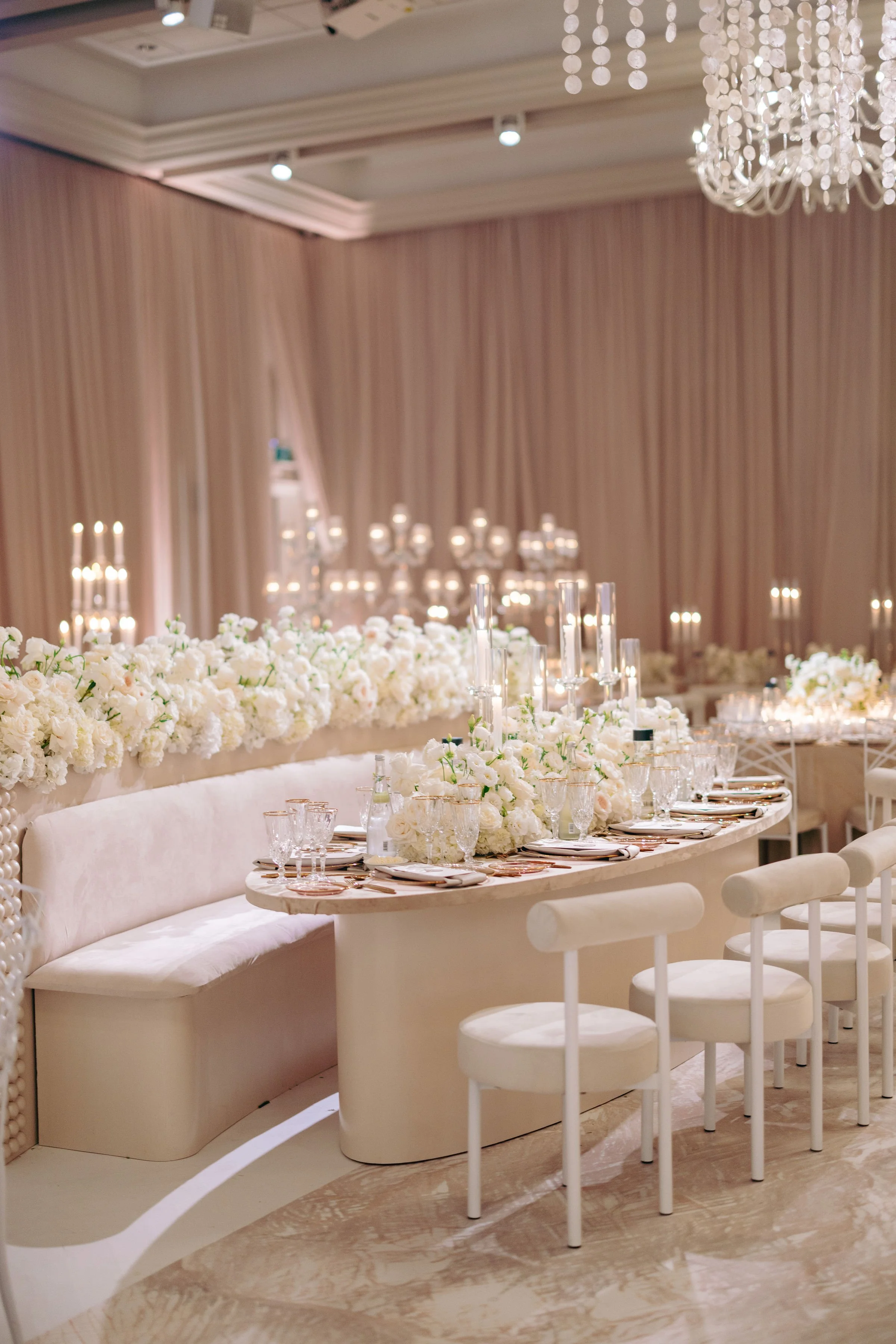 Elegant wedding reception table with white flowers, candles, glassware, and gold utensils in a luxurious decorated room.
