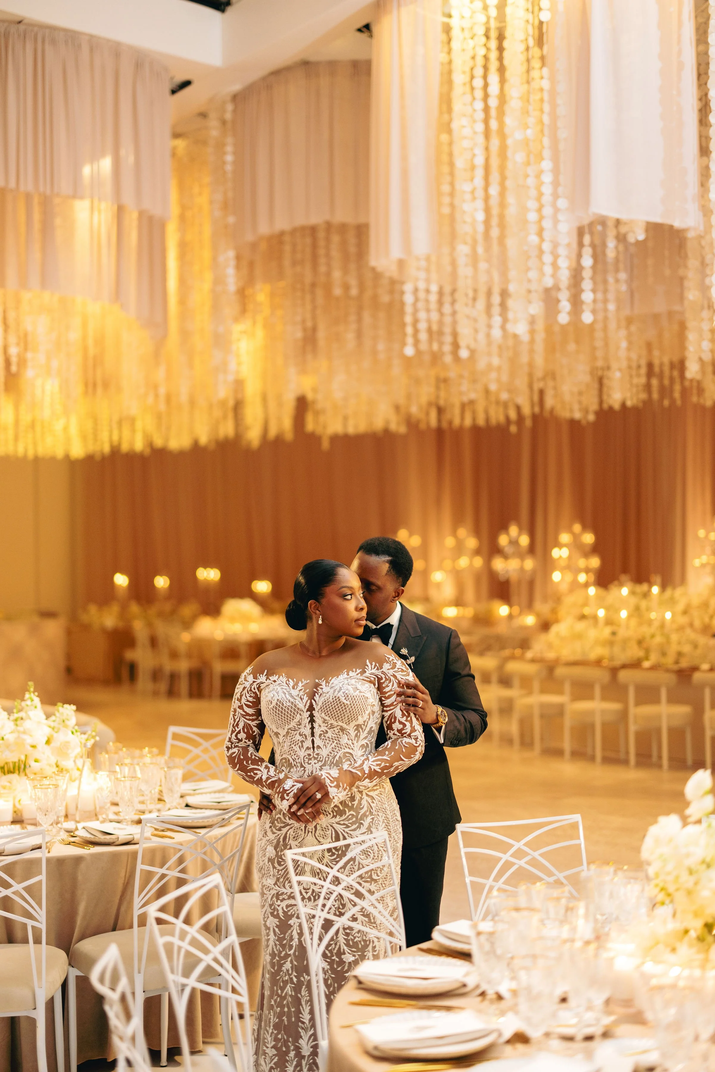 A newlywed couple in elegant wedding attire standing close together in a lavishly decorated reception hall with gold and white decor, surrounded by tables set for a celebration.