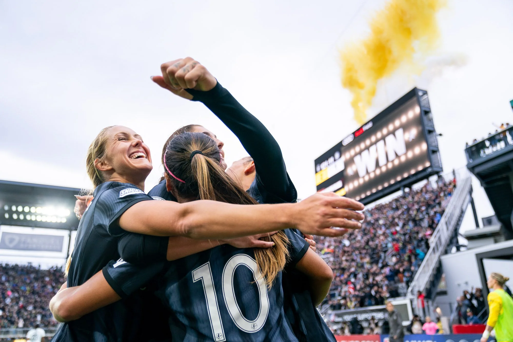 Washington Spirit forward Brittany Ratcliffe embraces her teammates with a smile immediately after the final whistle of the 2024 NWSL Playoff Quarterfinal victory over Bay FC at Audi Field. The word “WIN” is displayed on the stadium video board as ye