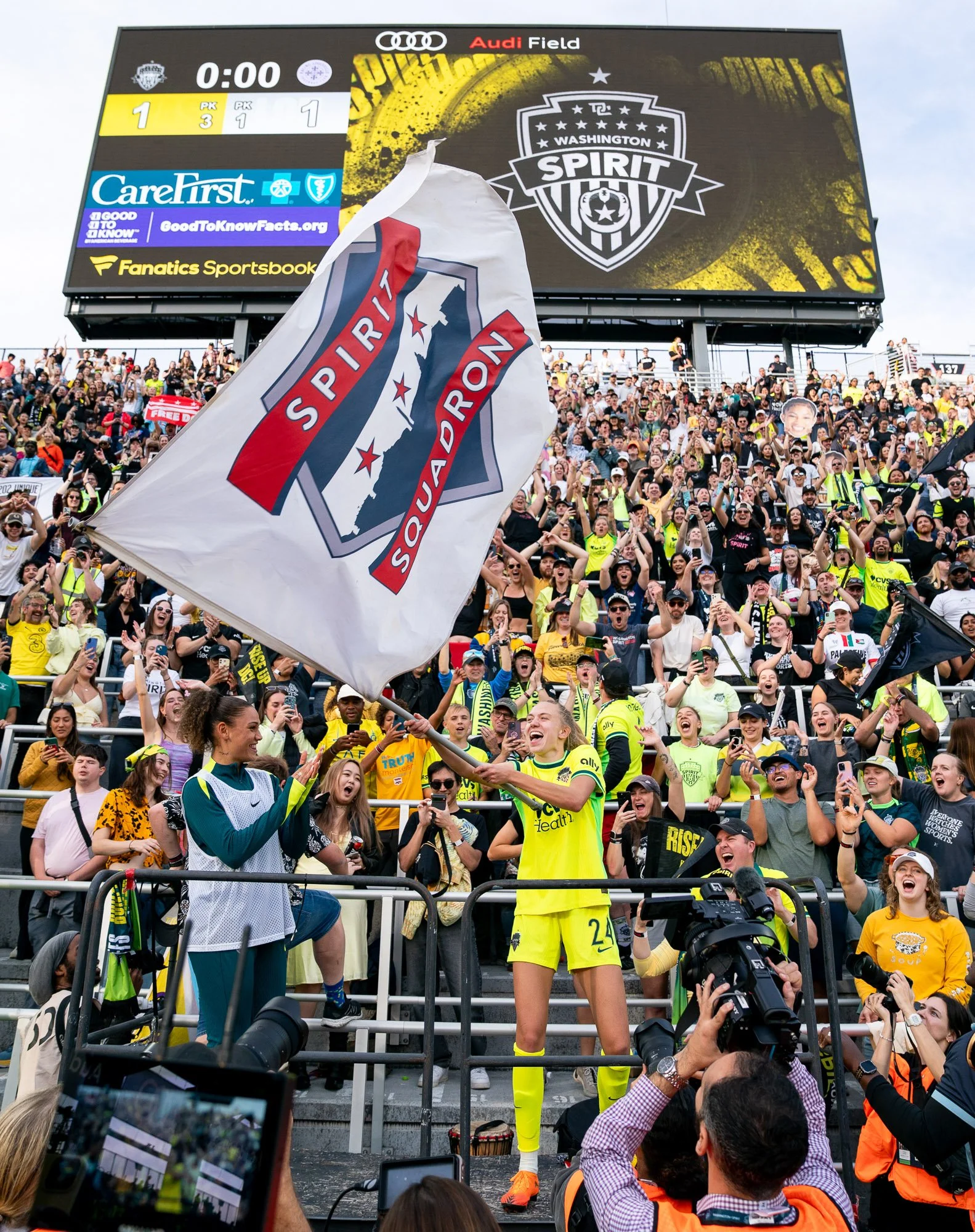 Washington Spirit defender Esme Morgan waves a Spirit Squadron flag toward the supporters section as teammate Trinity Rodman looks on following the Spirit’s 2025 NWSL Playoff Quarterfinal victory over Racing Louisville FC at Audi Field. Fans fill the