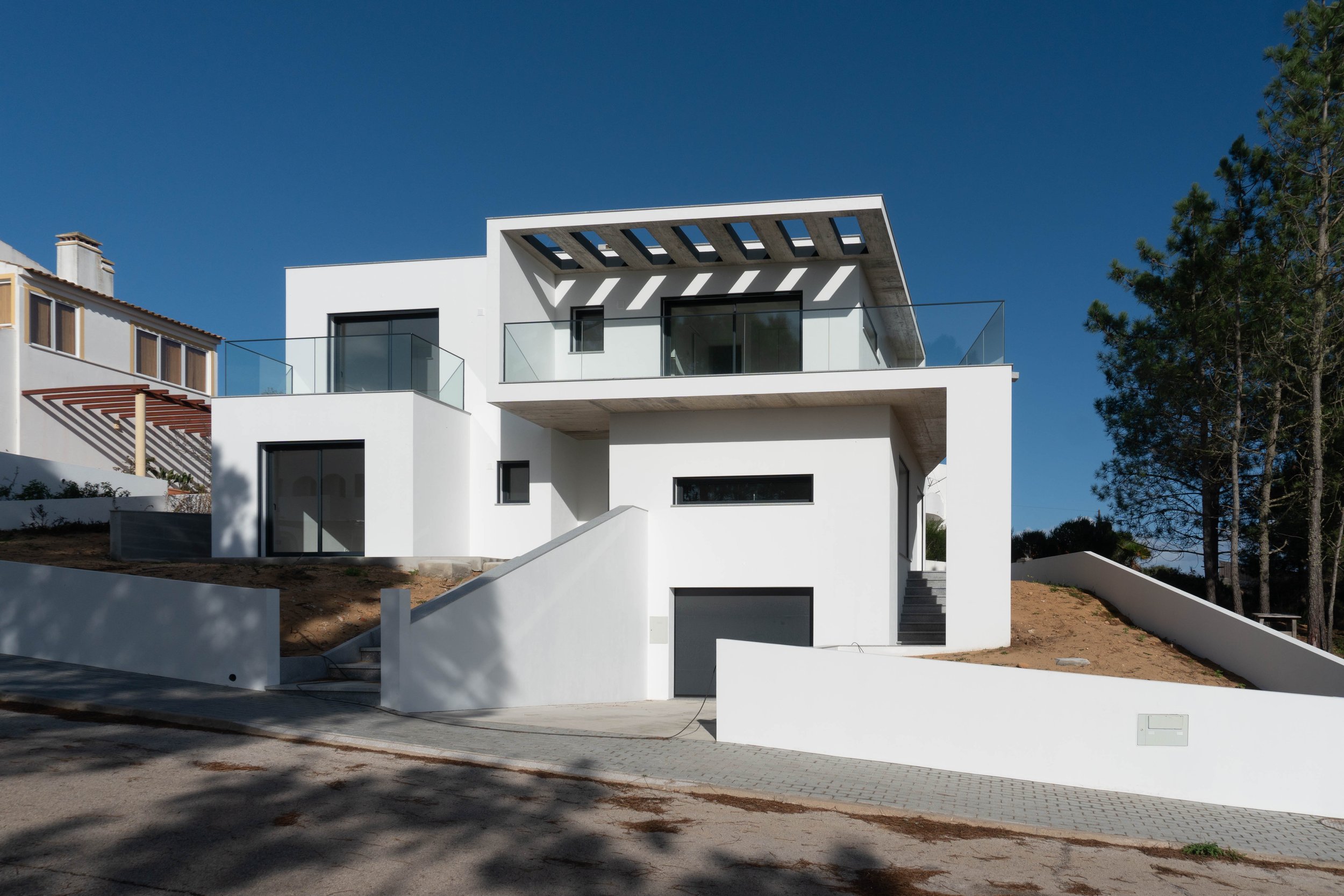 Casa moderna de dois andares com fachada branca, varandas de vidro e design arquitetônico minimalista. Árvore ao lado e céu azul ao fundo.