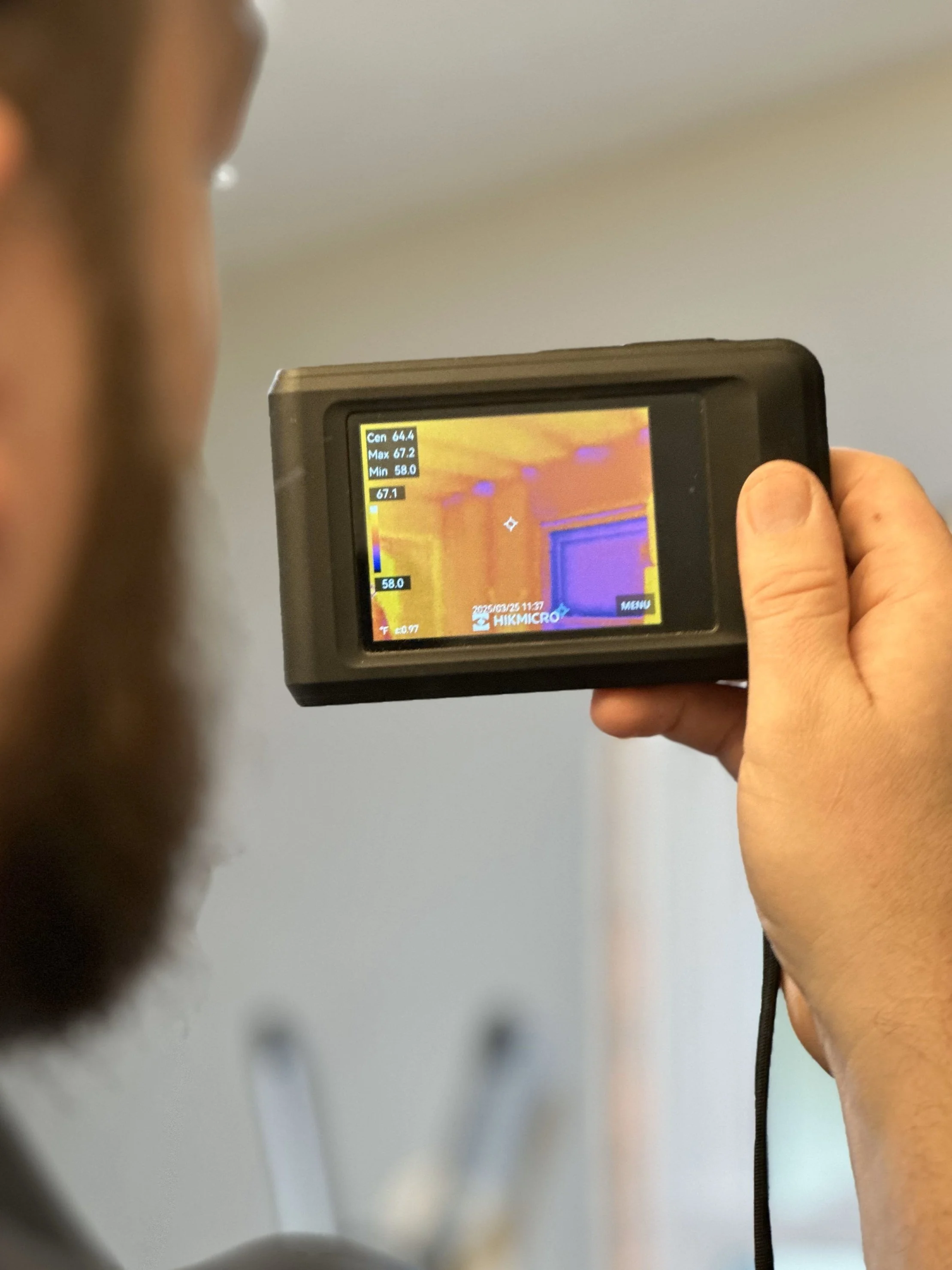 Person holding a thermal imaging camera, showing a thermal image of a room with warm and cool areas.