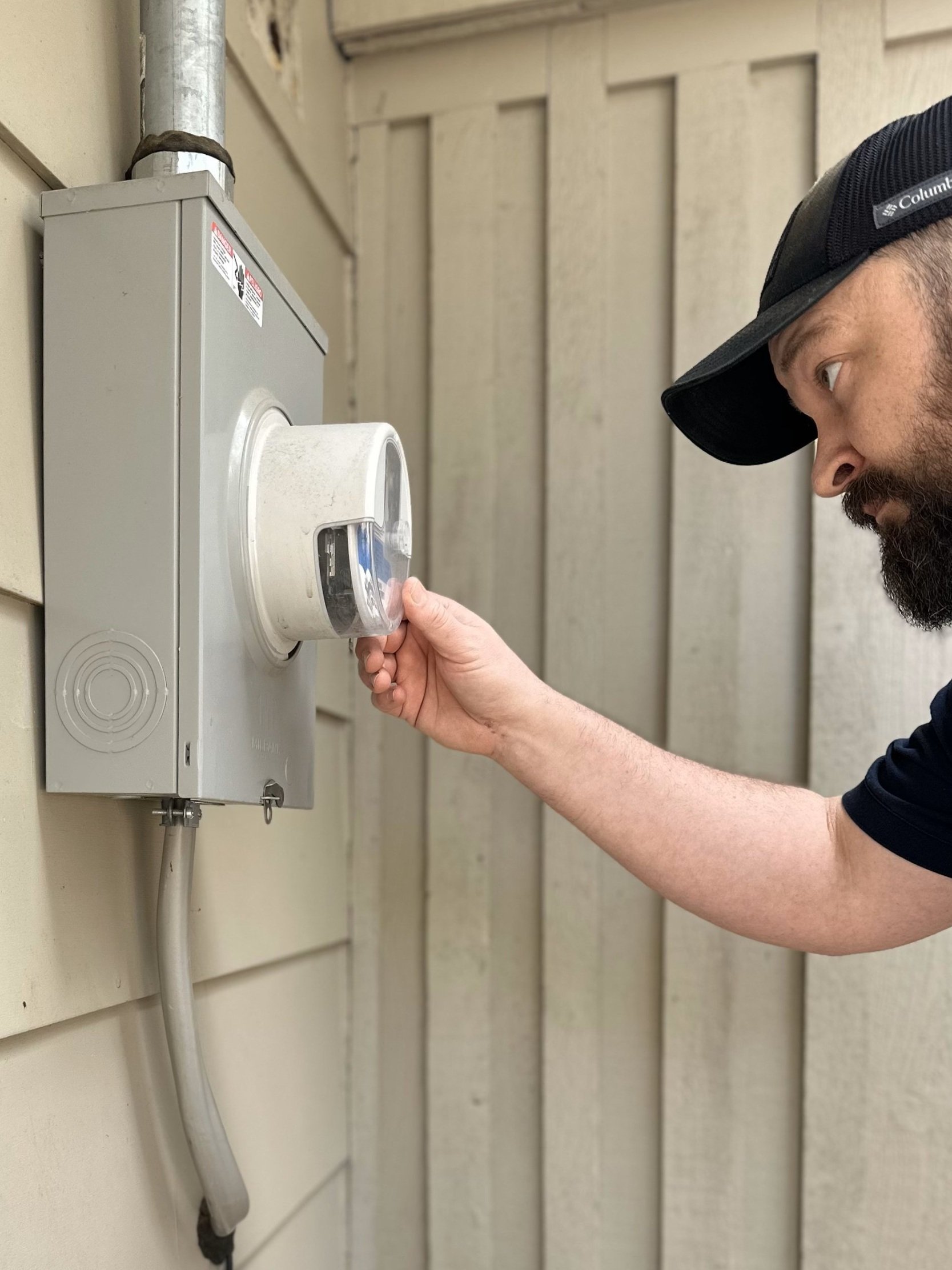 A man wearing a black Columbia cap and a navy shirt is plugging a device into an outdoor electrical outlet mounted on a beige siding wall.