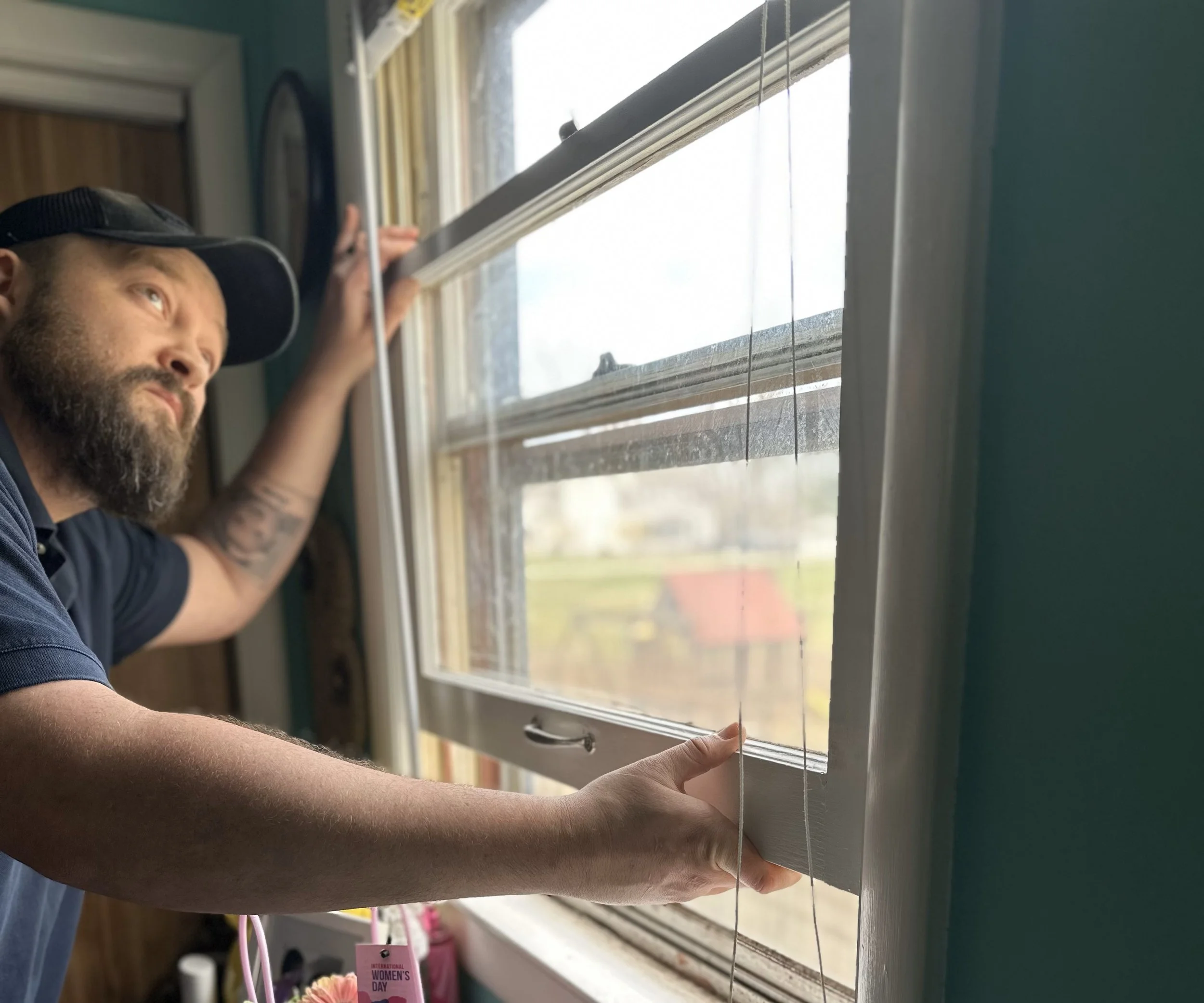 A man with a beard and a black cap is opening a window, with sunlight shining inside. The window has white frames and is slightly open, with outside scenery visible.