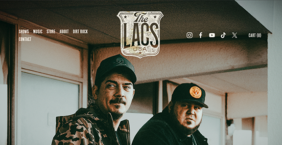 Two men sitting inside a music venue or bar, with one smiling and wearing a camouflage jacket and baseball cap, the other with a serious expression wearing a black hoodie and baseball cap with a logo. The background features a window and enclosed space.