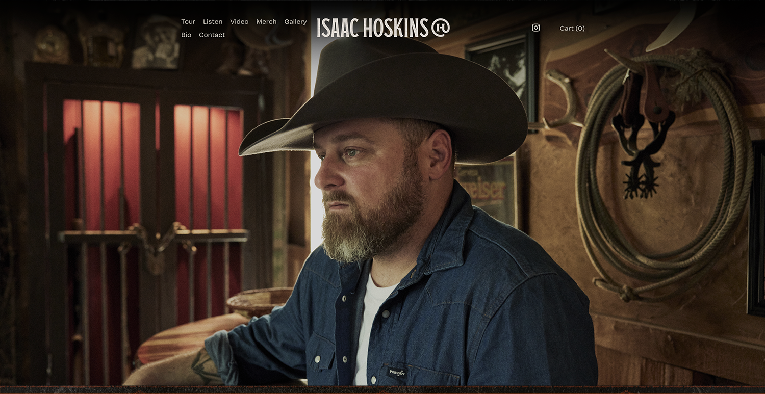 A man with a beard wearing a cowboy hat and denim shirt inside a rustic room with cowboy gear on the wall.